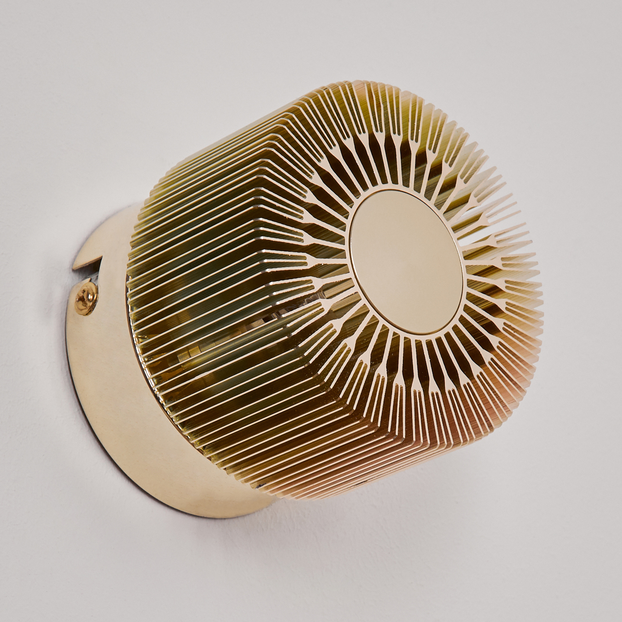 Eine goldene zylindrische Wandlampe mit geriffelter Oberfläche und kreisförmiger Mitte, an einer weißen Wand befestigt.