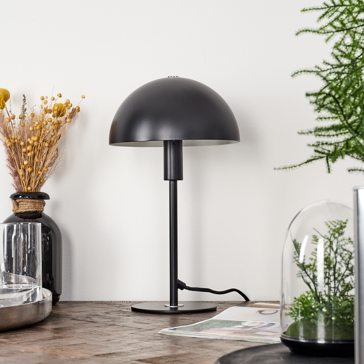 Eine schwarze Schreibtischlampe mit einem Kuppelschirm steht auf einem Holztisch. Eine Vase mit Trockenblumen ist links. Rechts eine kleine Pflanze in einer Glaskuppel.