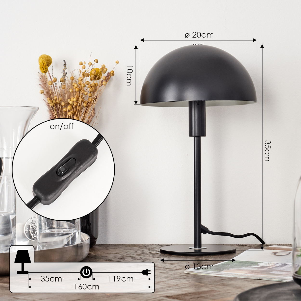 Eine schwarze Schreibtischlampe mit einem kuppelförmigen Schirm, die Abmessungen sind angegeben. Ein Ein/Aus-Schalter und Trockenblumen sind ebenfalls zu sehen.