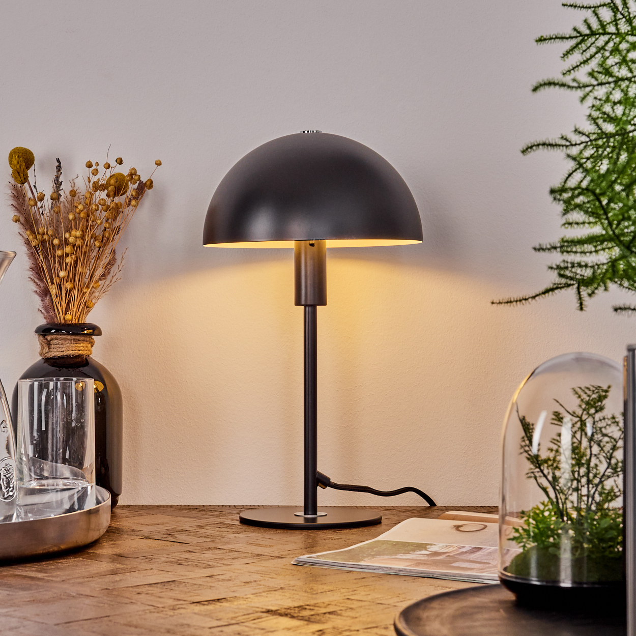 Eine schwarze Lampe mit einem gewölbten Schirm steht auf einem Holztisch. Eine Vase mit Trockenblumen, ein Glas und eine Zeitschrift sind ebenfalls auf dem Tisch.