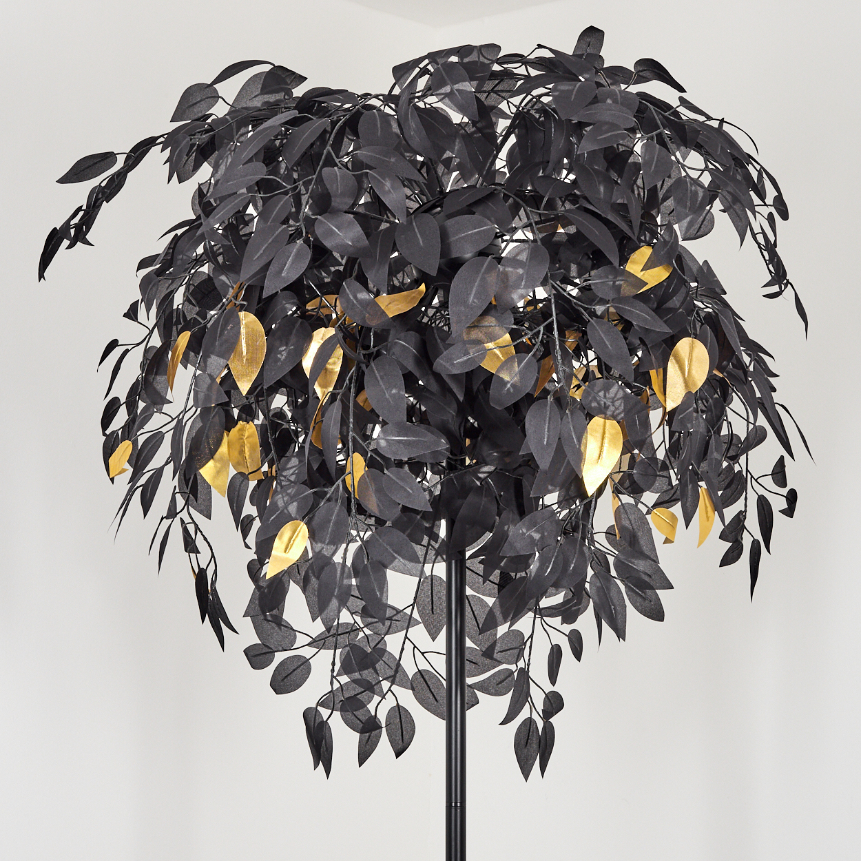 Ein schwarzer Kunstbaum mit goldenen Blättern. Er hat eine schwarze Metallstange und steht vor einer weißen Wand.