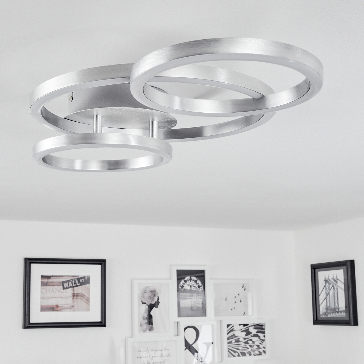 Eine moderne silberne Deckenleuchte mit drei sich überlappenden Ringen. Darunter befinden sich gerahmte Bilder an einer weißen Wand.