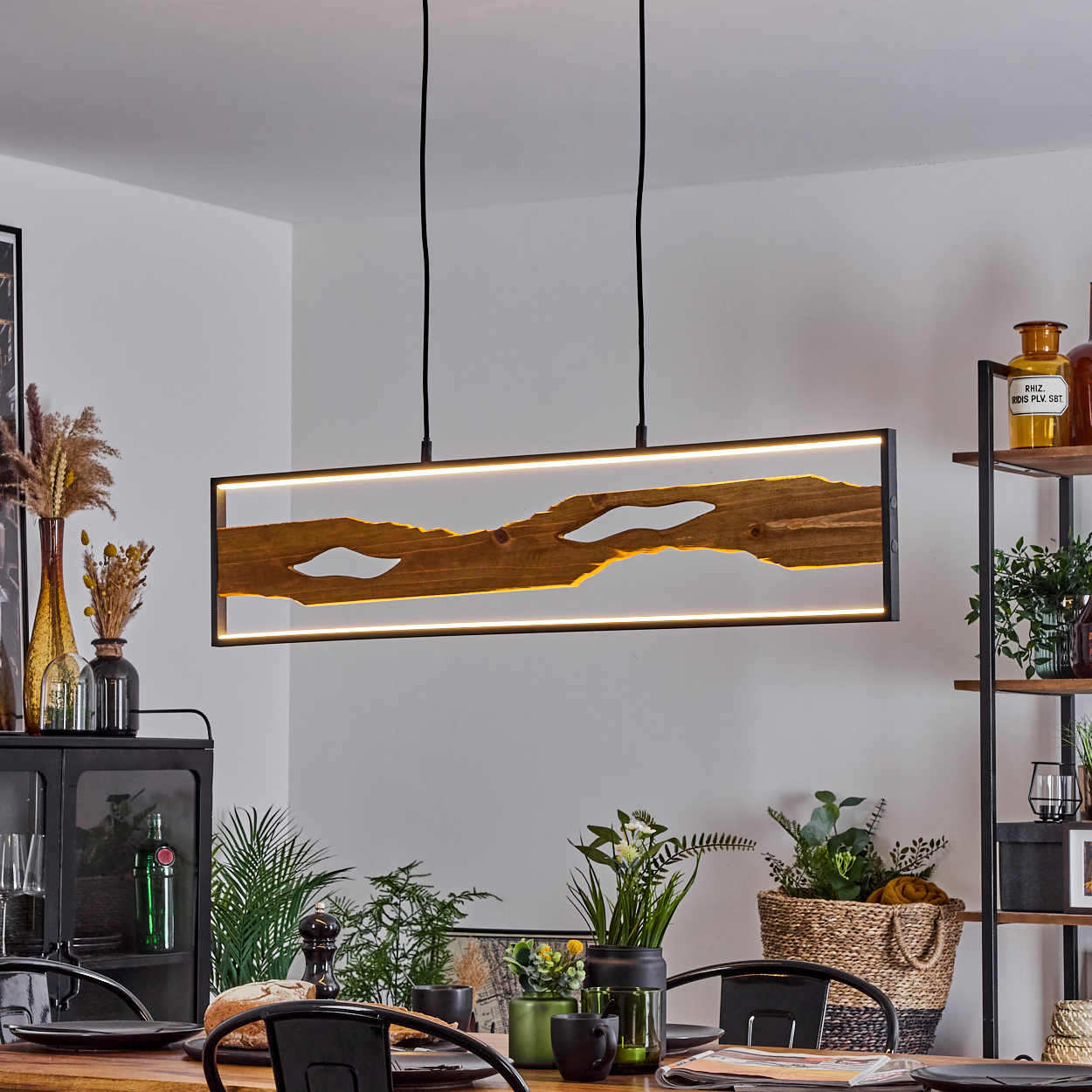 Ein Esszimmer mit Holztisch, schwarzen Stühlen und einer rechteckigen Holzlampe. Im Hintergrund Regale und Pflanzen.