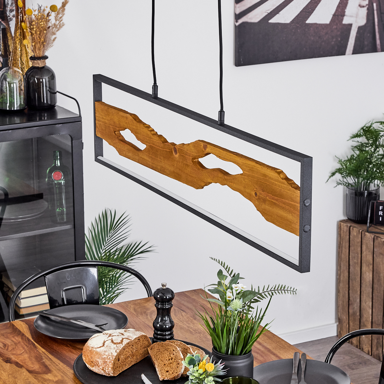 Essszene mit Holztisch, Brot, Tellern und einer modernen Holz-Metall-Lampe. Hintergrund Pflanzen und Dekoration.