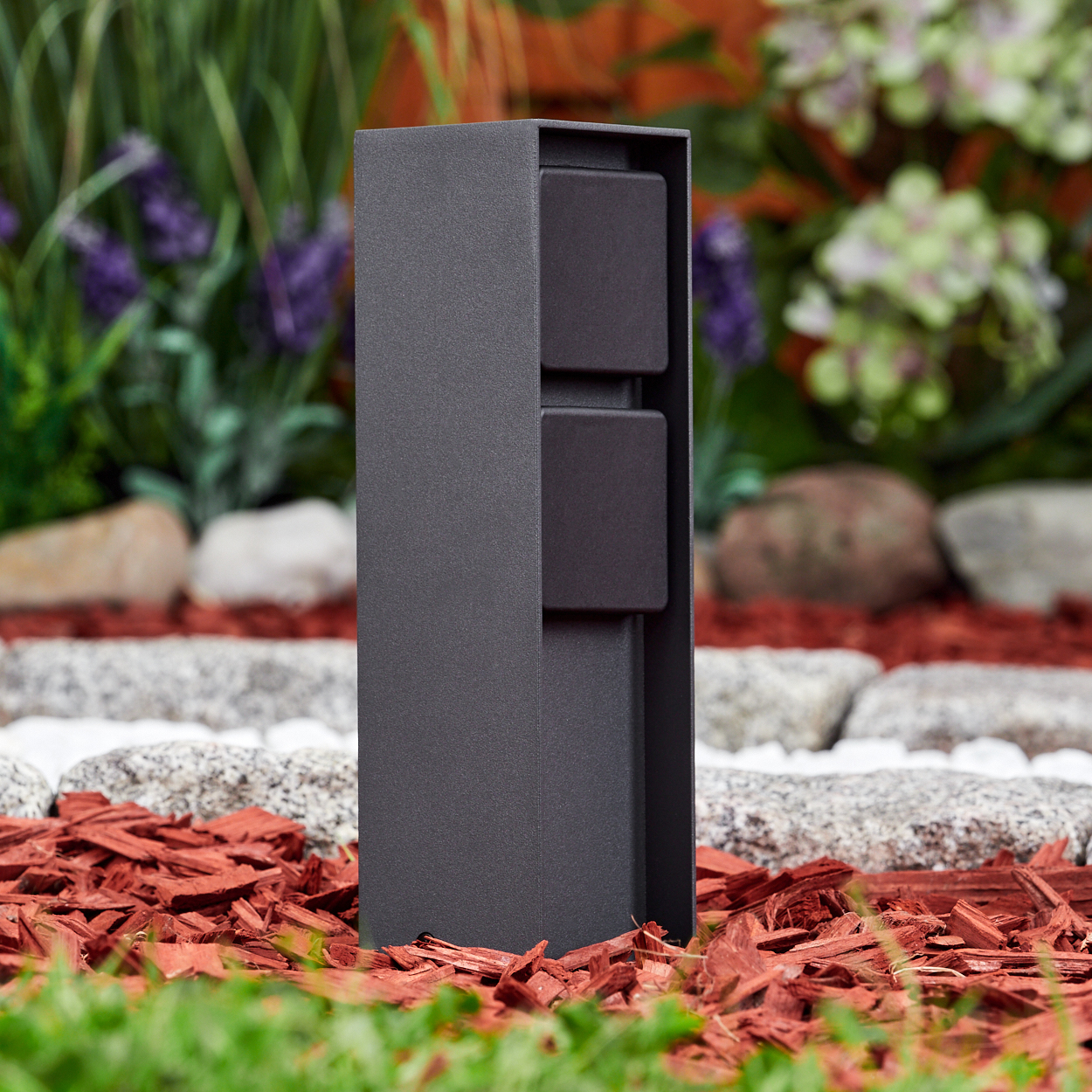 Eine dunkelgraue Außenleuchte in einem Garten. Die Leuchte hat zwei dunkle quadratische Elemente. Der Hintergrund hat Steine und Pflanzen.