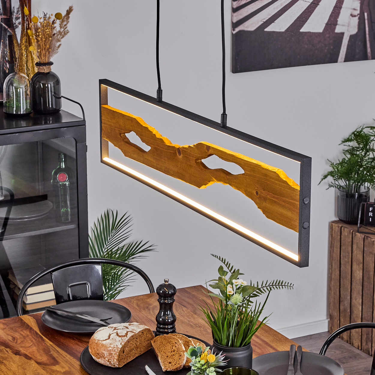 Esszimmer mit Holztisch, schwarzer Lampe, Brot, Tellern und Pflanzen. Eine Holzplatte mit Licht.