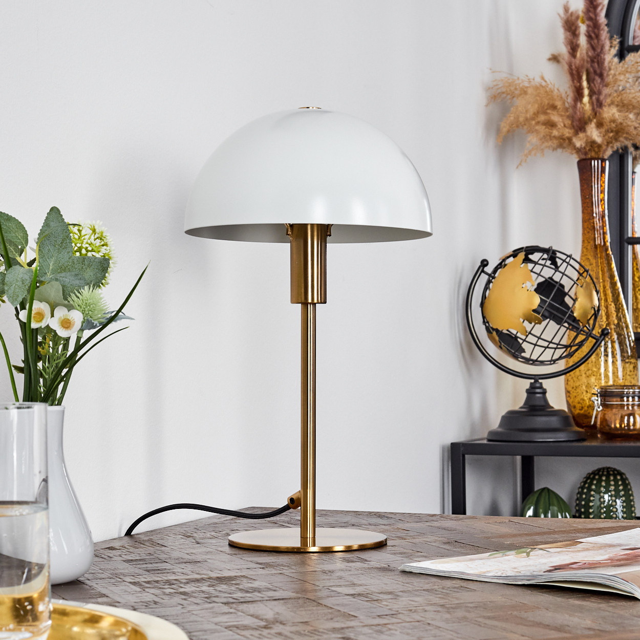 Eine weiße Kuppellampe mit goldenem Stiel steht auf einem Holztisch. Weitere Artikel sind Globus, Vasen, Blumen und eine Zeitschrift.