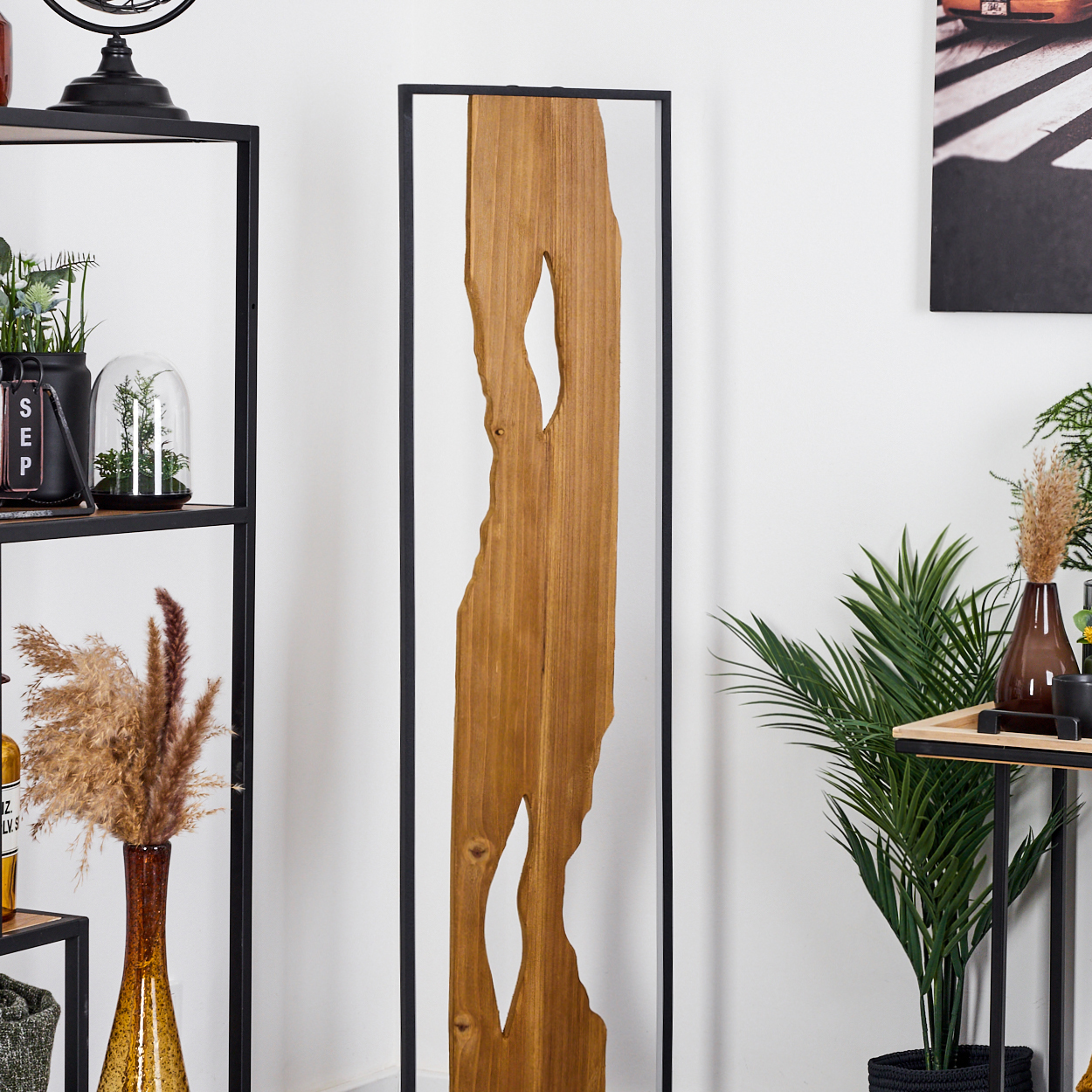 Ein Holzkunstwerk mit Ausschnitten steht vor einer weißen Wand. Regale mit Pflanzen und ein schwarzer Rahmen sind zu sehen.