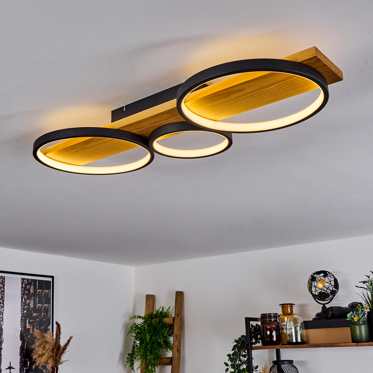 Eine Deckenleuchte mit drei kreisförmigen Lichtern. Sie ist an einem Holzbrett befestigt. Der Hintergrund zeigt einen Raum mit Dekor.