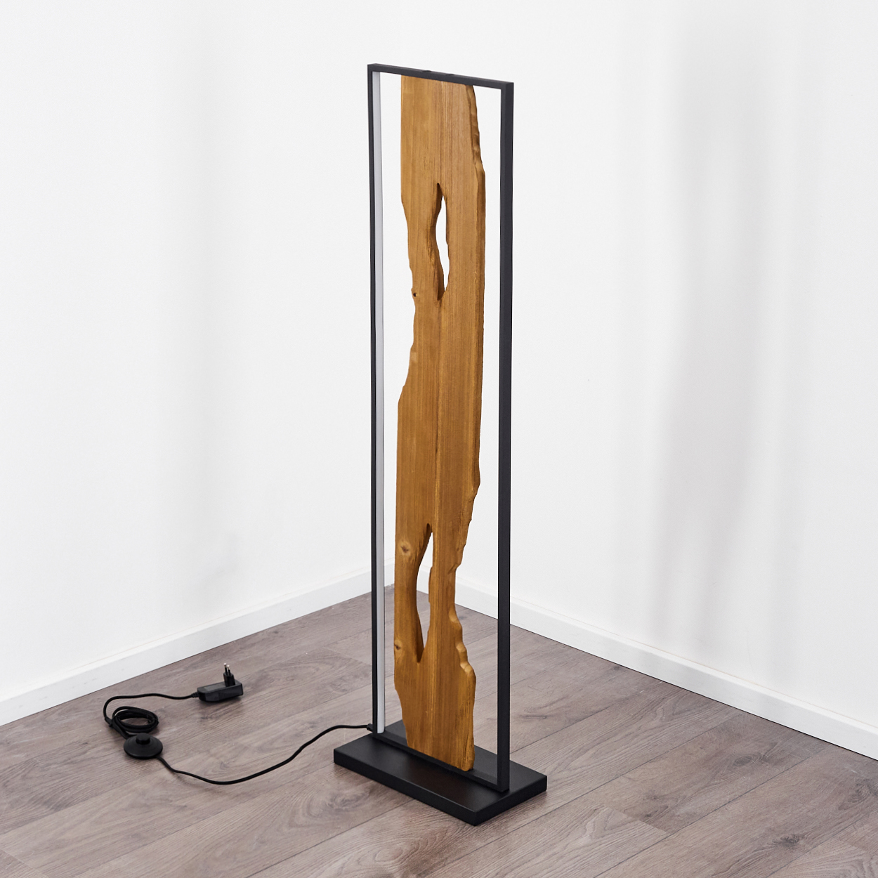Eine Holzlampe steht vor einer weißen Wand. Die Lampe hat einen rechteckigen schwarzen Rahmen und eine Holzplatte mit Aussparungen.