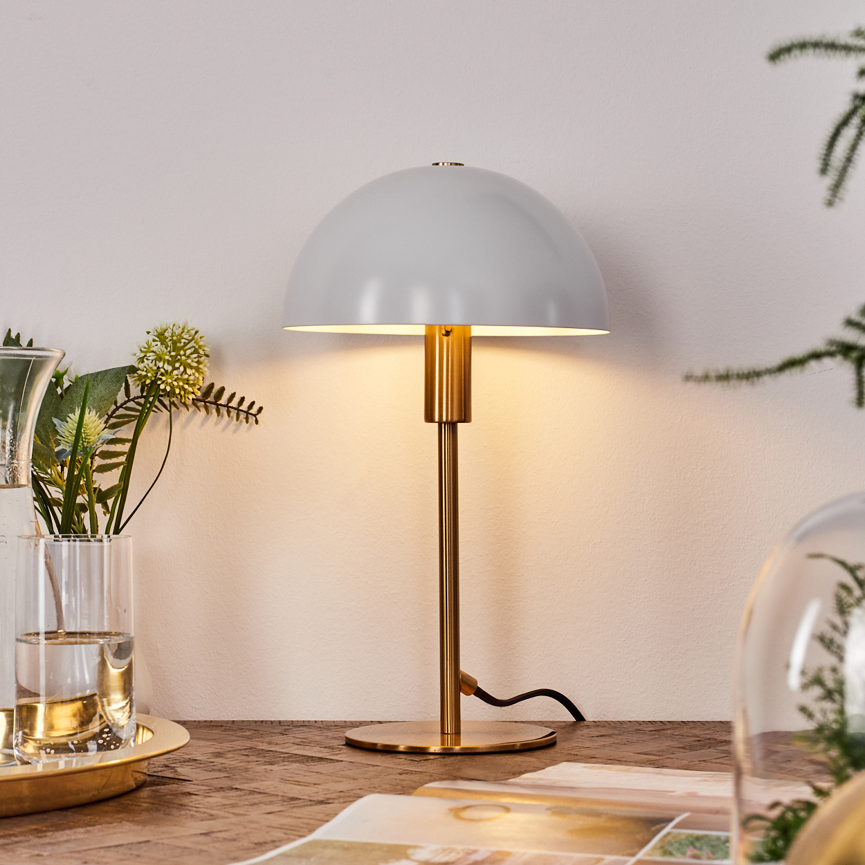 Eine graue Kuppellampe mit goldenem Fuß und Stiel steht auf einem Holztisch. Links ist eine Glasvase mit Blumen.
