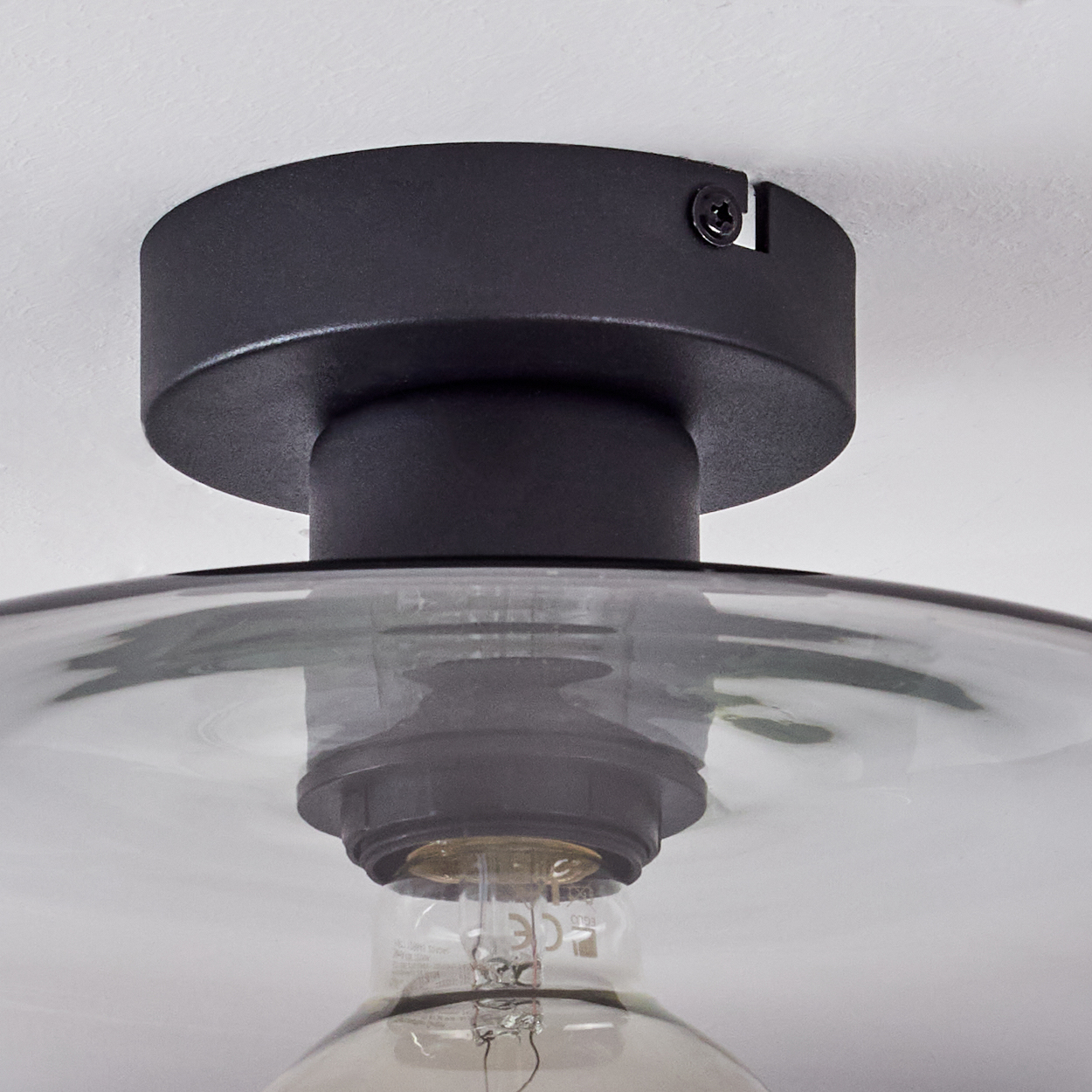Eine schwarze Deckenleuchte. Sie hat einen runden Sockel, der an der Decke befestigt ist. Darunter befindet sich ein Glasschirm. Die Glühbirne ist sichtbar.