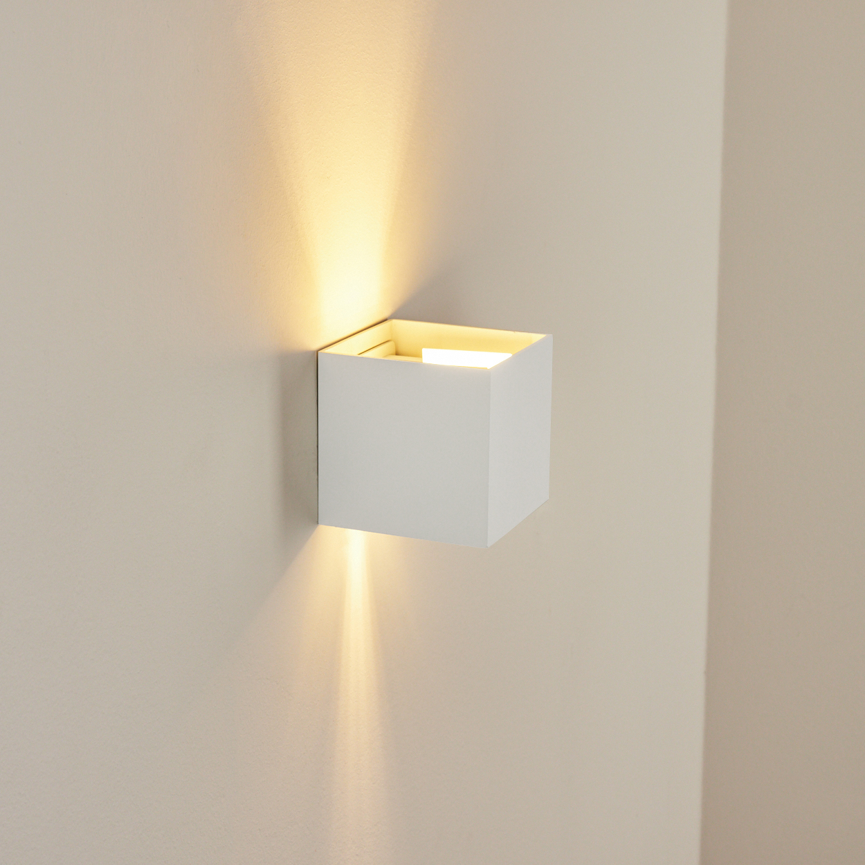 Eine weiße, würfelförmige Wandleuchte strahlt warmes Licht aus. Die Lampe ist an einer beigefarbenen Wand montiert.