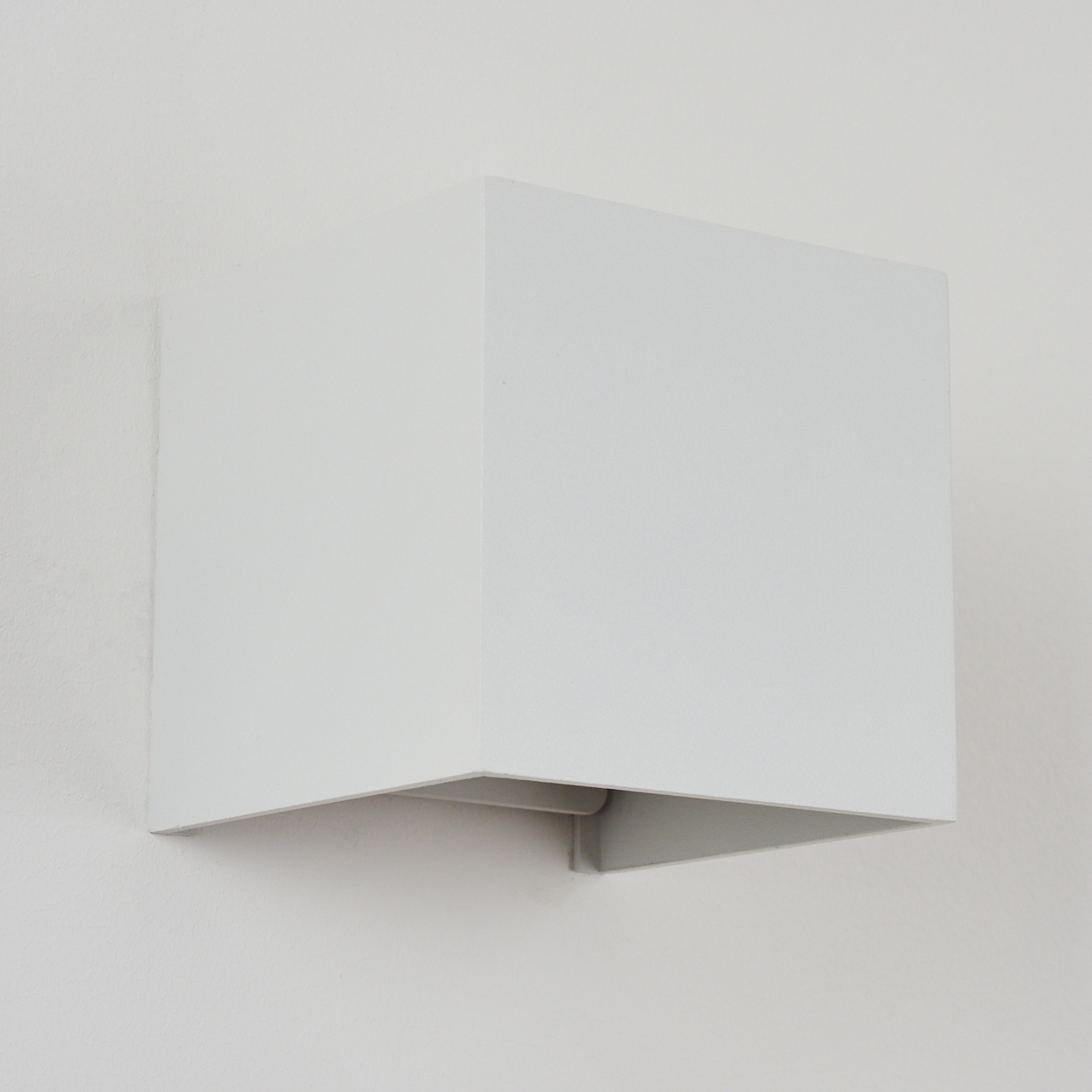 Eine weiße, würfelförmige Wandleuchte ist an einer weißen Wand angebracht. Die Lampe hat eine offene Unterseite.