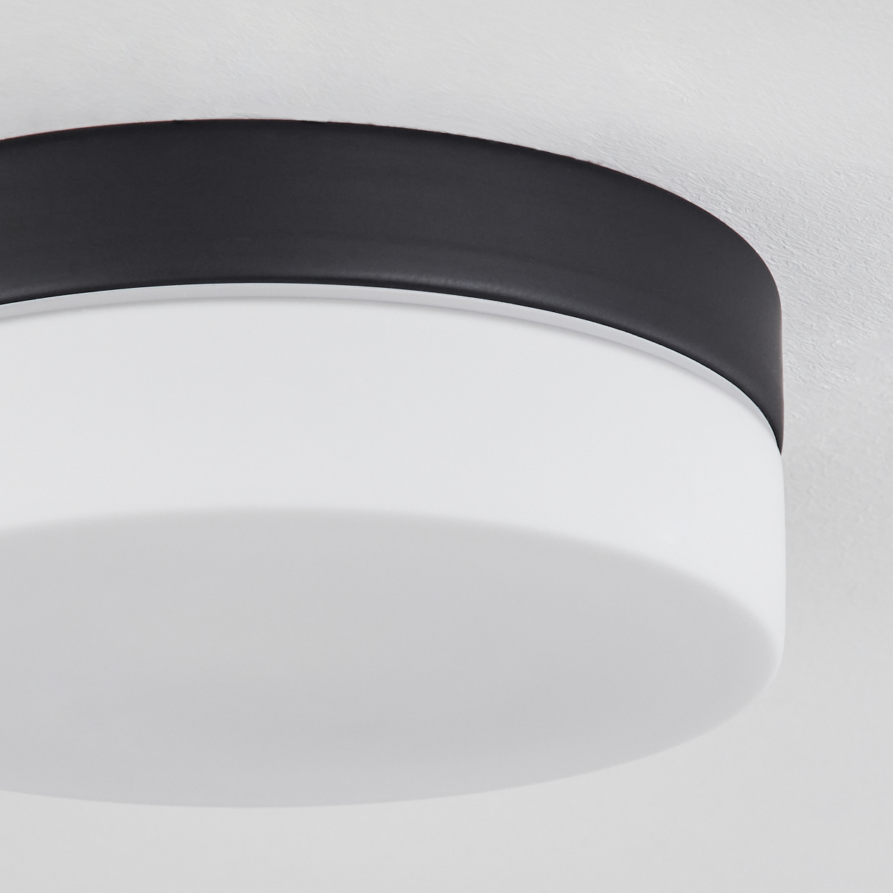 Nahaufnahme einer Deckenlampe. Sie hat einen weißen, abgerundeten Sockel mit schwarzem Rand. Der Hintergrund ist eine weiße Decke.