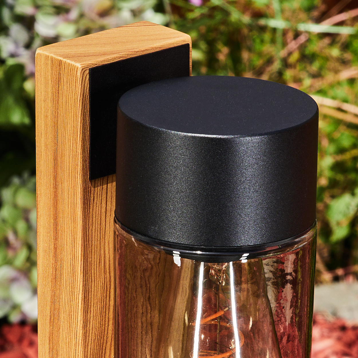 Ein Holzpfosten mit einer schwarzen Lampe. Die Lampe hat eine Glasabdeckung und eine sichtbare Glühbirne. Im Hintergrund sind grüne Pflanzen.