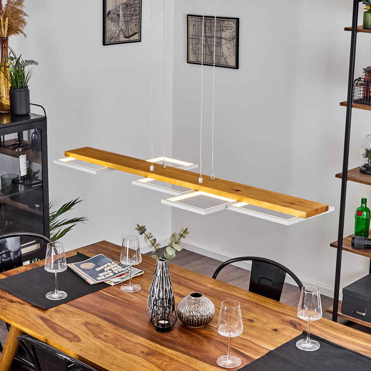 Ein Esszimmer mit Holztisch, Kronleuchter und Dekoration. Schwarze Stühle und ein Regal mit Gegenständen sind sichtbar.