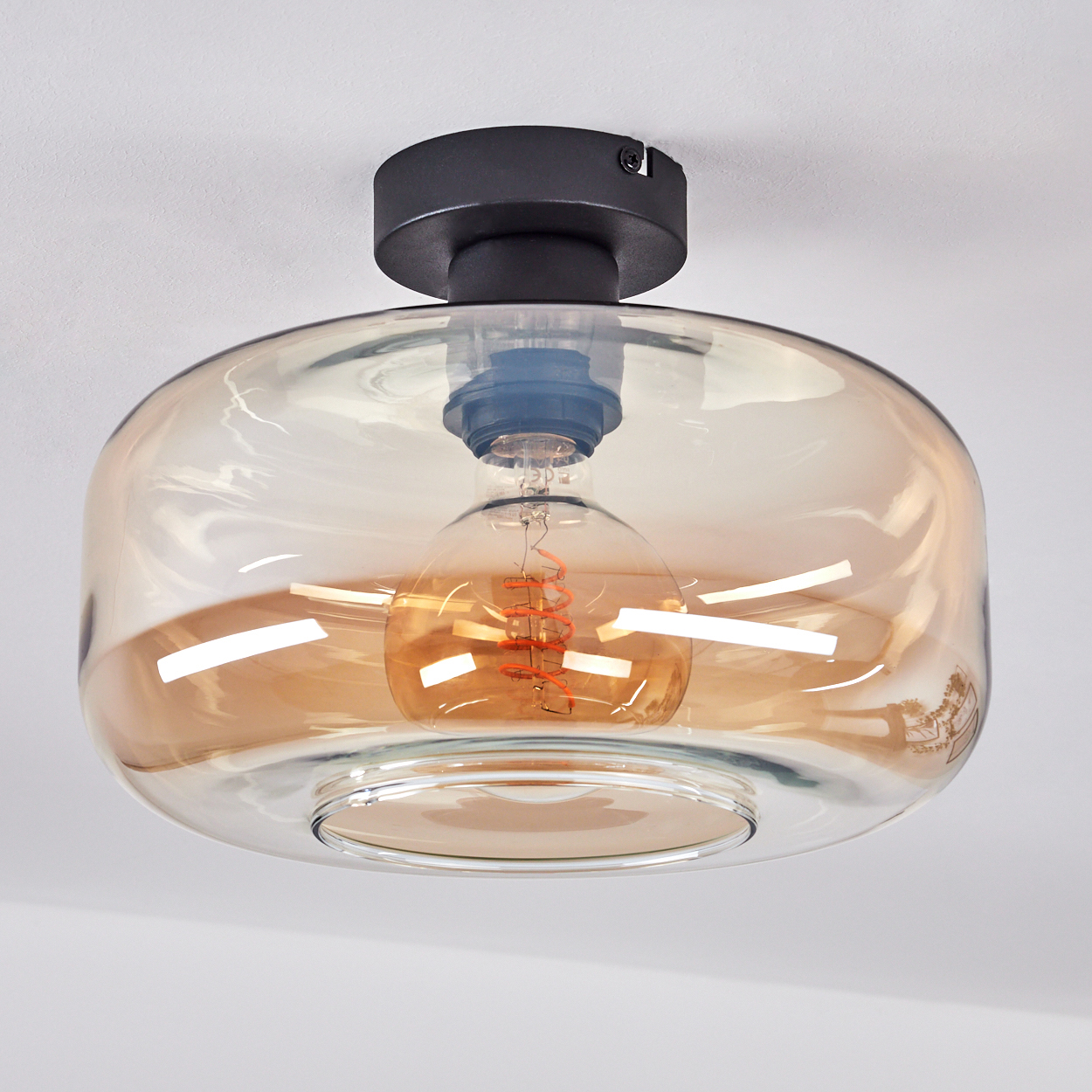Eine Deckenlampe mit bernsteinfarbenem Glasschirm. Im Inneren befindet sich eine sichtbare Glühbirne. Die Lampe ist an einer weißen Decke montiert.