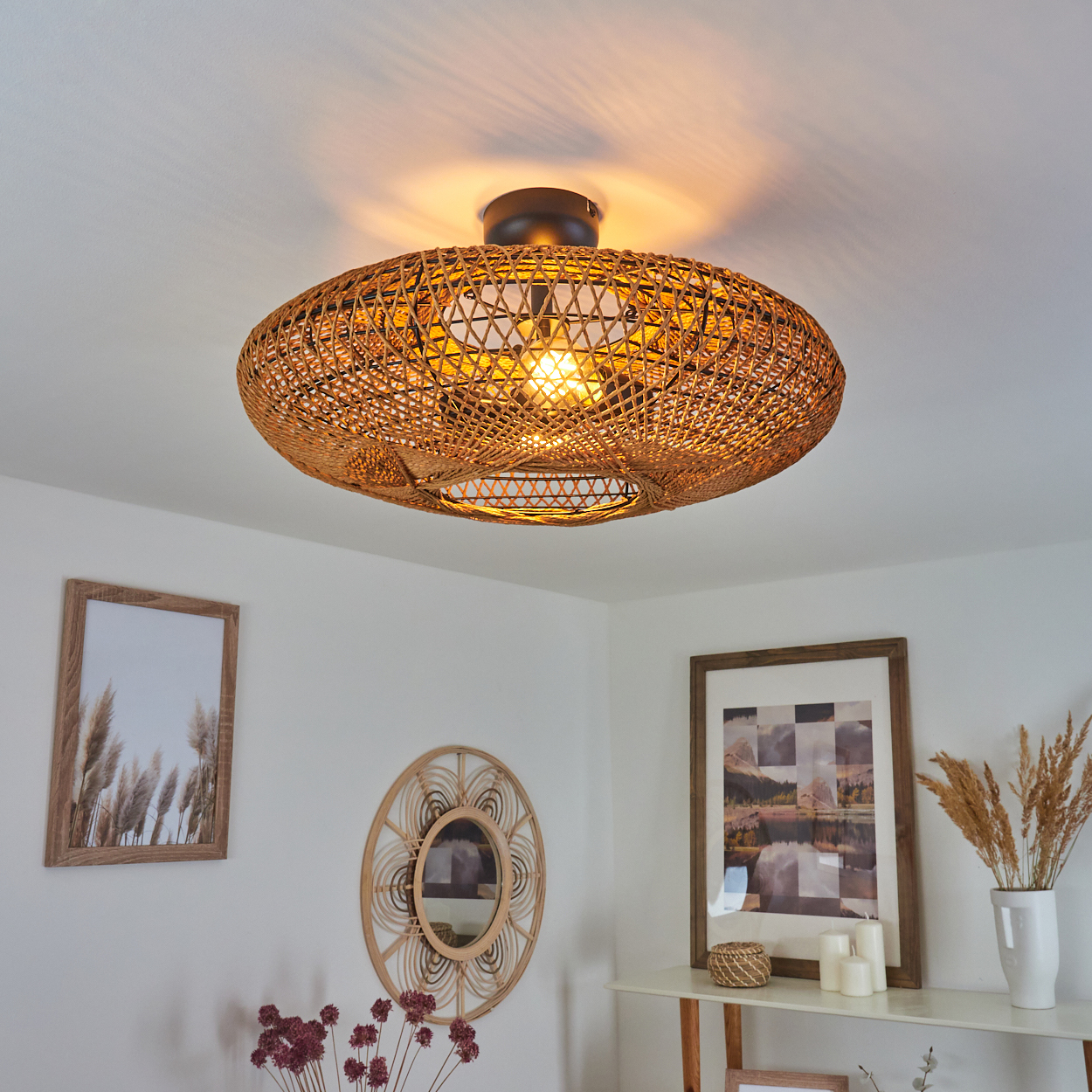 Eine Deckenlampe aus geflochtenem Rattan beleuchtet einen Raum mit Kunstwerken und Dekoration. Weiße Wände, gerahmte Bilder und ein runder Spiegel sind sichtbar.