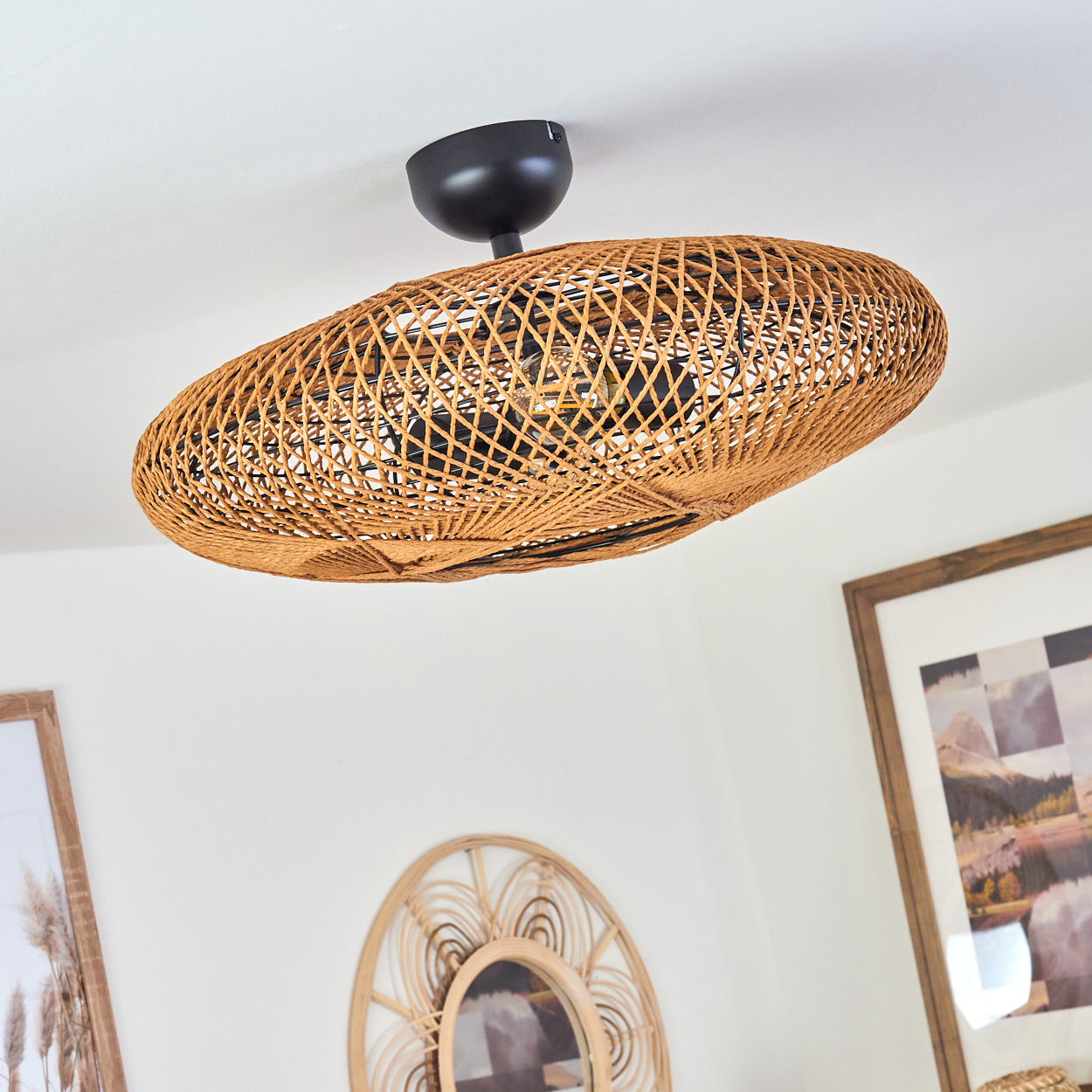 Ein Deckenventilator aus Korbgeflecht hängt von einer weißen Decke. Darunter schmücken ein runder Spiegel und gerahmte Bilder die Wand.