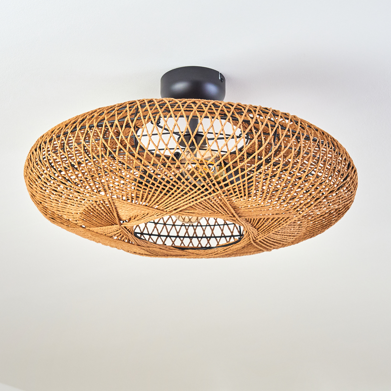 Eine gewebte Rattan-Leuchte hängt an einer weißen Decke. Die Leuchte ist oval mit einem schwarzen Sockel.