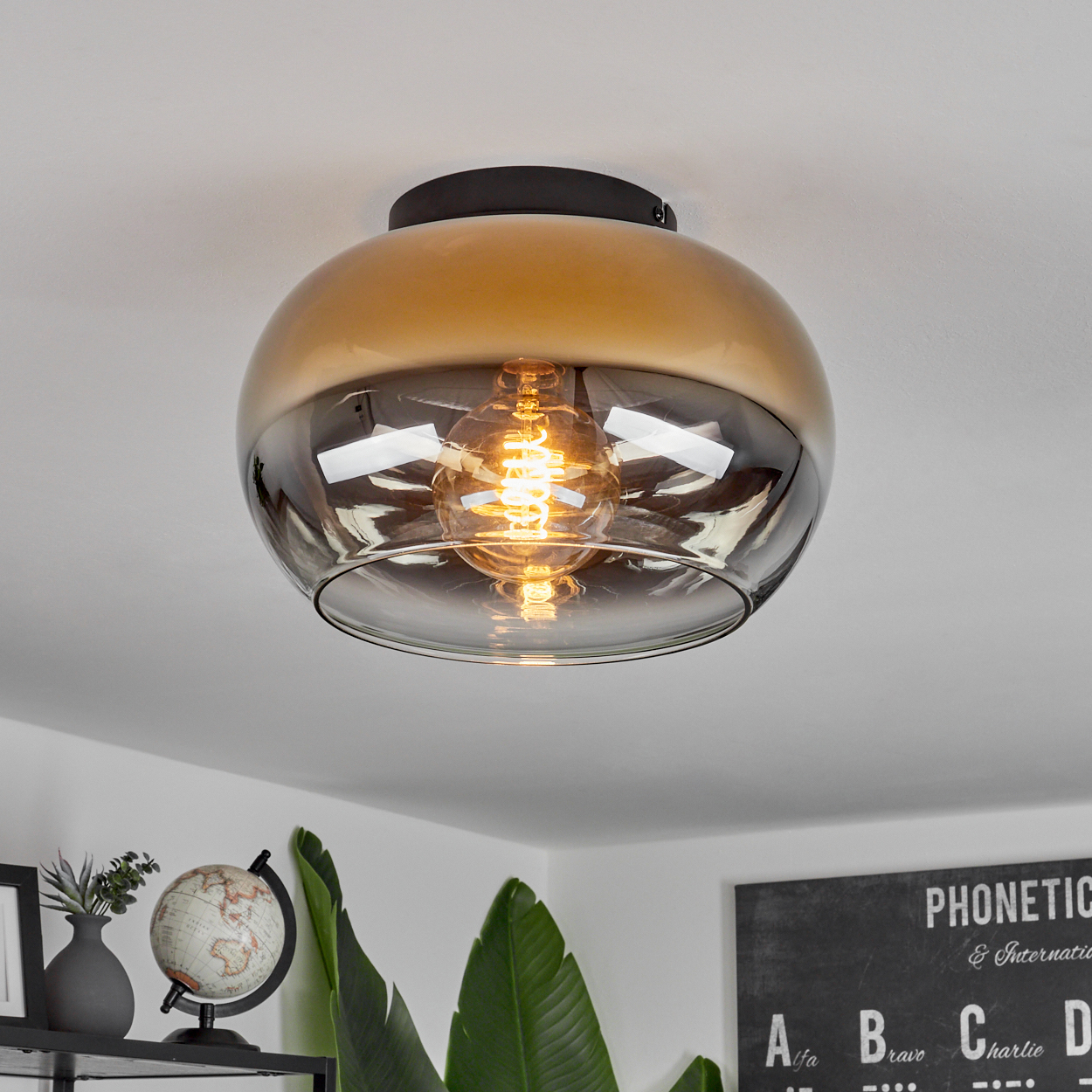 Eine moderne Deckenleuchte mit goldbraunem Glasschirm. Im Inneren ist eine glühende Glühbirne zu sehen. Im Hintergrund sind ein Globus und Pflanzen zu sehen.