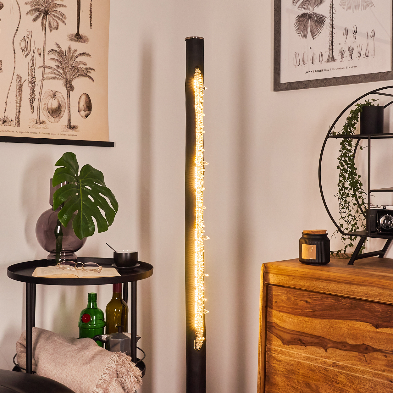 Ein Raum mit einer schwarzen Stehlampe mit Lichtern, einem Beistelltisch mit Gegenständen, einem Holzschrank und botanischen Drucken an der Wand.