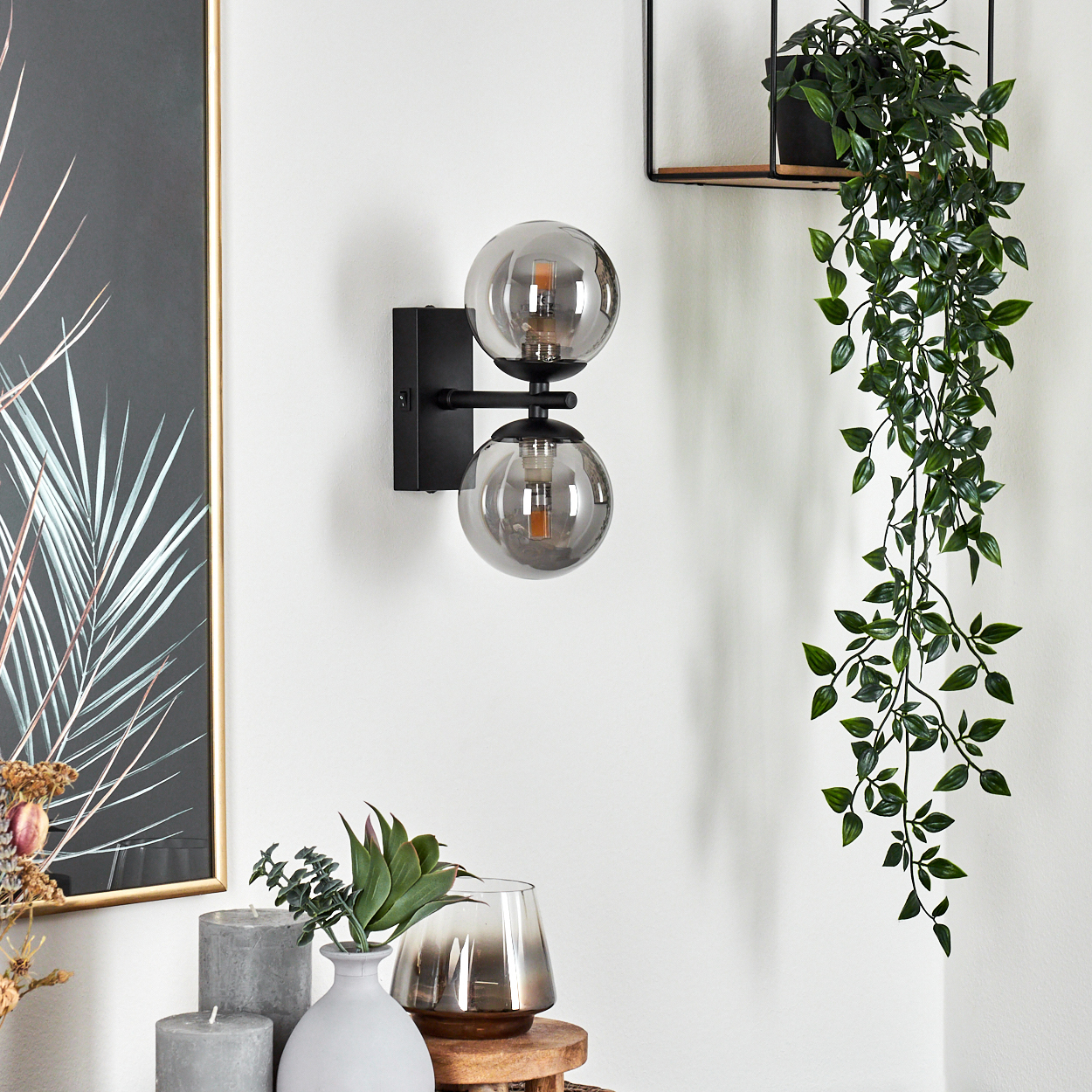 Eine Wandlampe mit zwei Glaskugelschirmen, schwarzem Rahmen und einer Hängepflanze. Weitere Dekorationen sind Kerzen und Vasen.