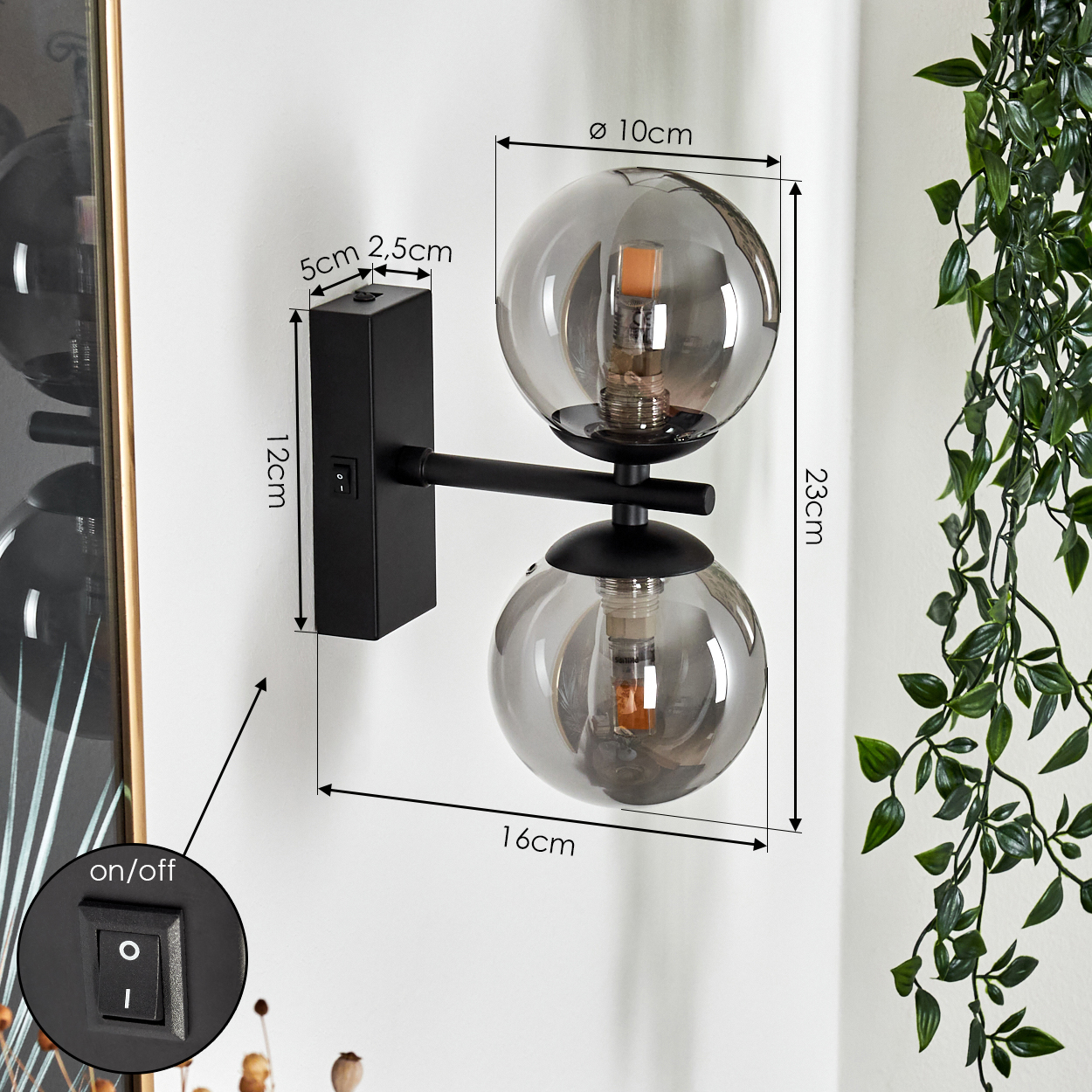 Schwarze Wandlampe mit zwei Glaskugeln. Maße sind angegeben. Grüne Pflanze im Hintergrund. Ein/Aus-Schalter an der Wand.