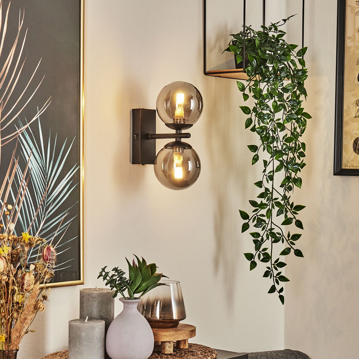 Eine Wand mit Lampe, Pflanze und Dekoration. Die Lampe hat zwei Kugeln. Eine Rankpflanze hängt. Es gibt Kerzen und eine Vase.