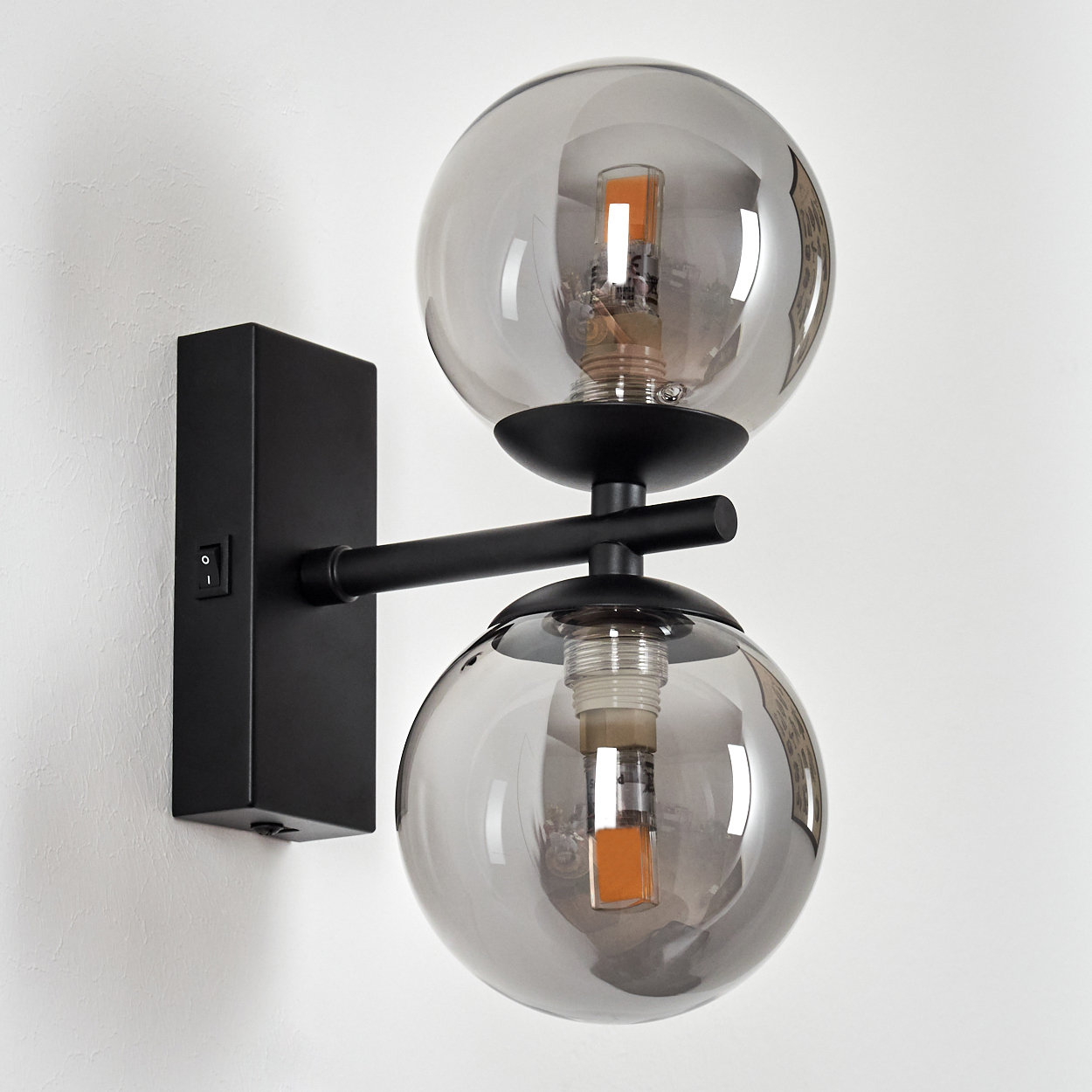 Eine schwarze Wandlampe mit zwei Glaskugelschirmen. Sie ist an einer weißen Wand mit einem Ein/Aus-Schalter befestigt.