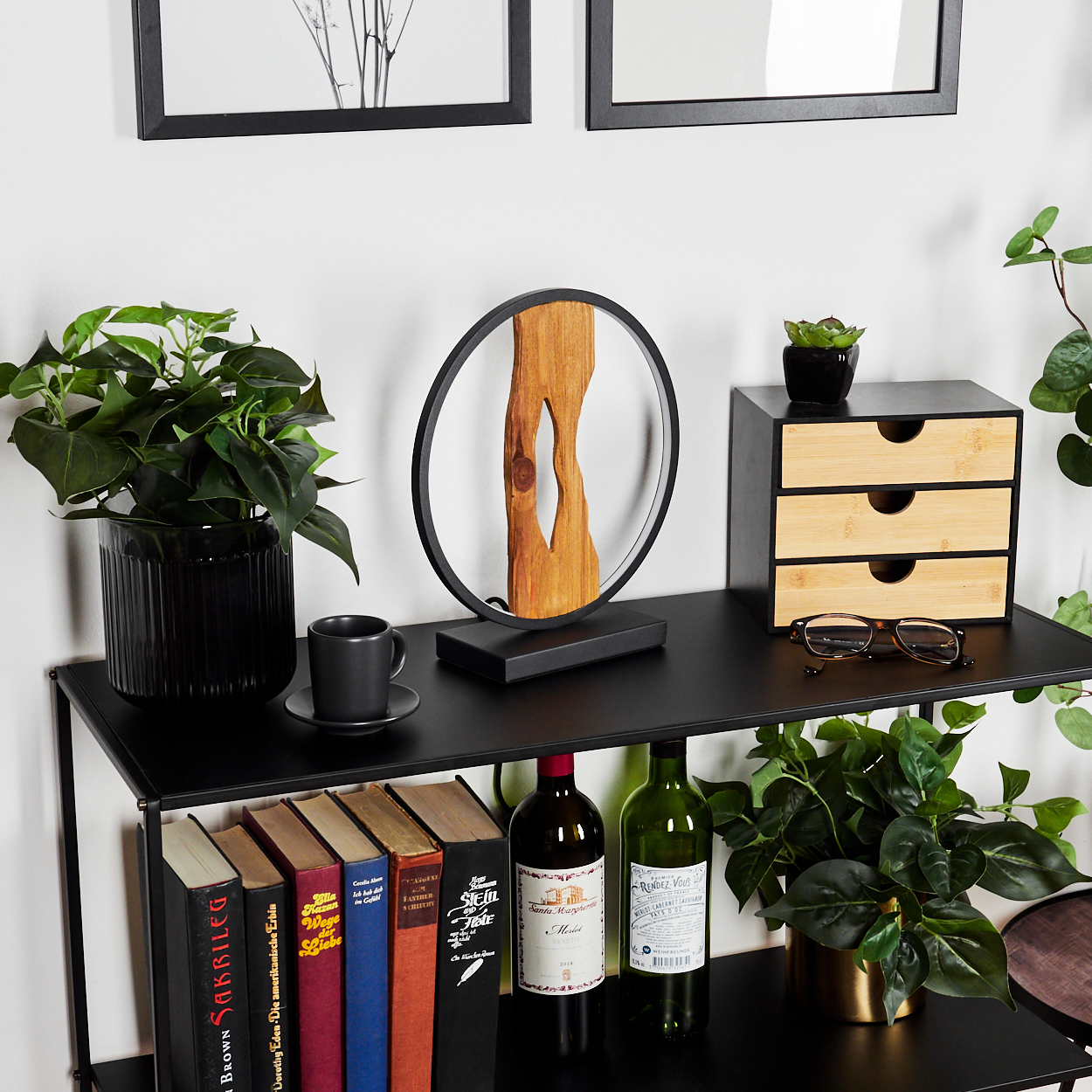 Ein schwarzes Regal mit Pflanzen, Büchern, Weinflaschen, einer Holzlampe, einer kleinen Schublade und einer Tasse.