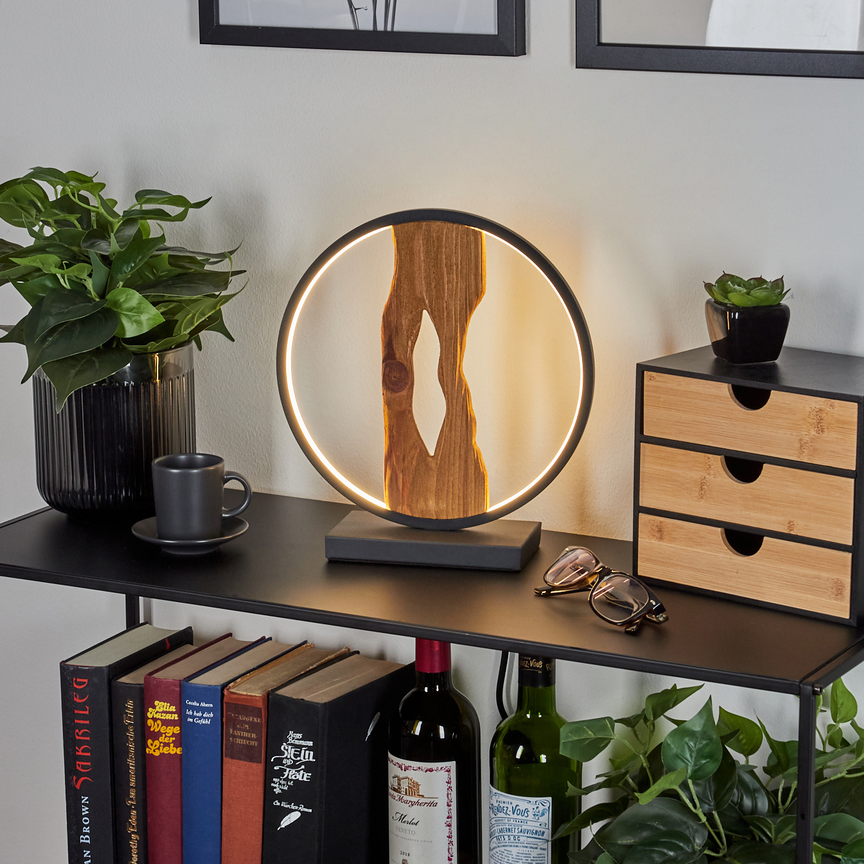 Eine kreisförmige Lampe mit Holzmittelstück steht auf einem Regal mit Büchern, Wein, einer Tasse und Pflanzen.