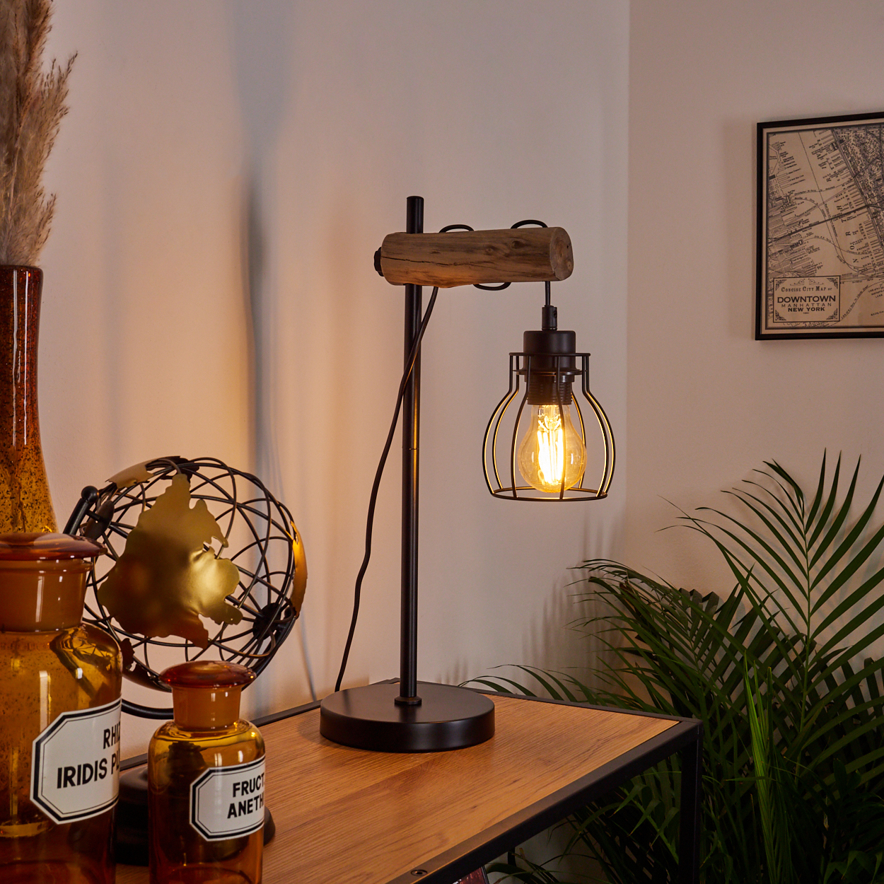 Eine Lampe mit Holz- und schwarzem Metalldesign steht auf einem Holztisch, mit anderen Gegenständen drumherum. An der Wand hängt eine Karte.