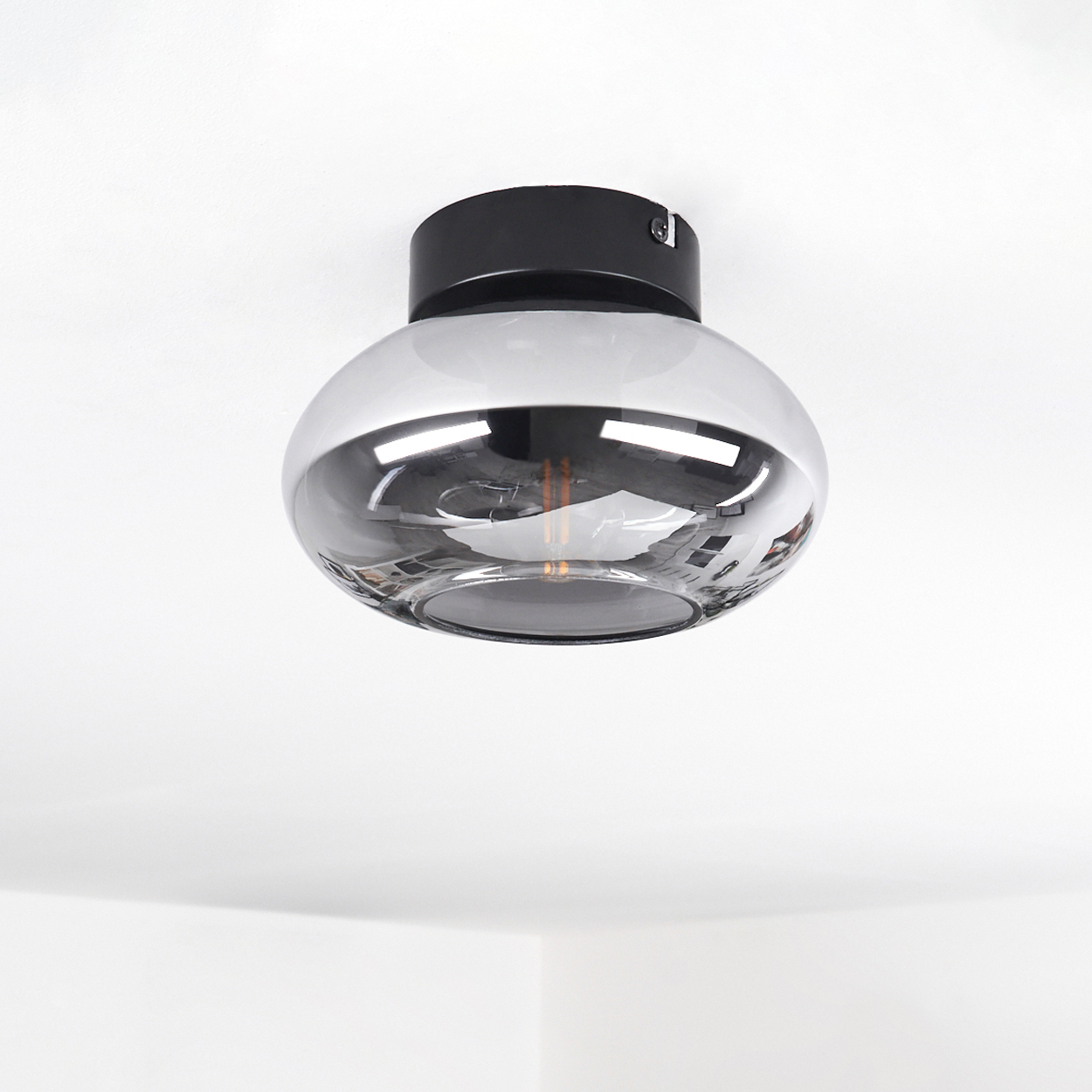 Eine Deckenlampe mit schwarzem Sockel und grauem, kugelförmigem Glasschirm. Die Lampe ist mittig an einer weißen Decke angebracht.