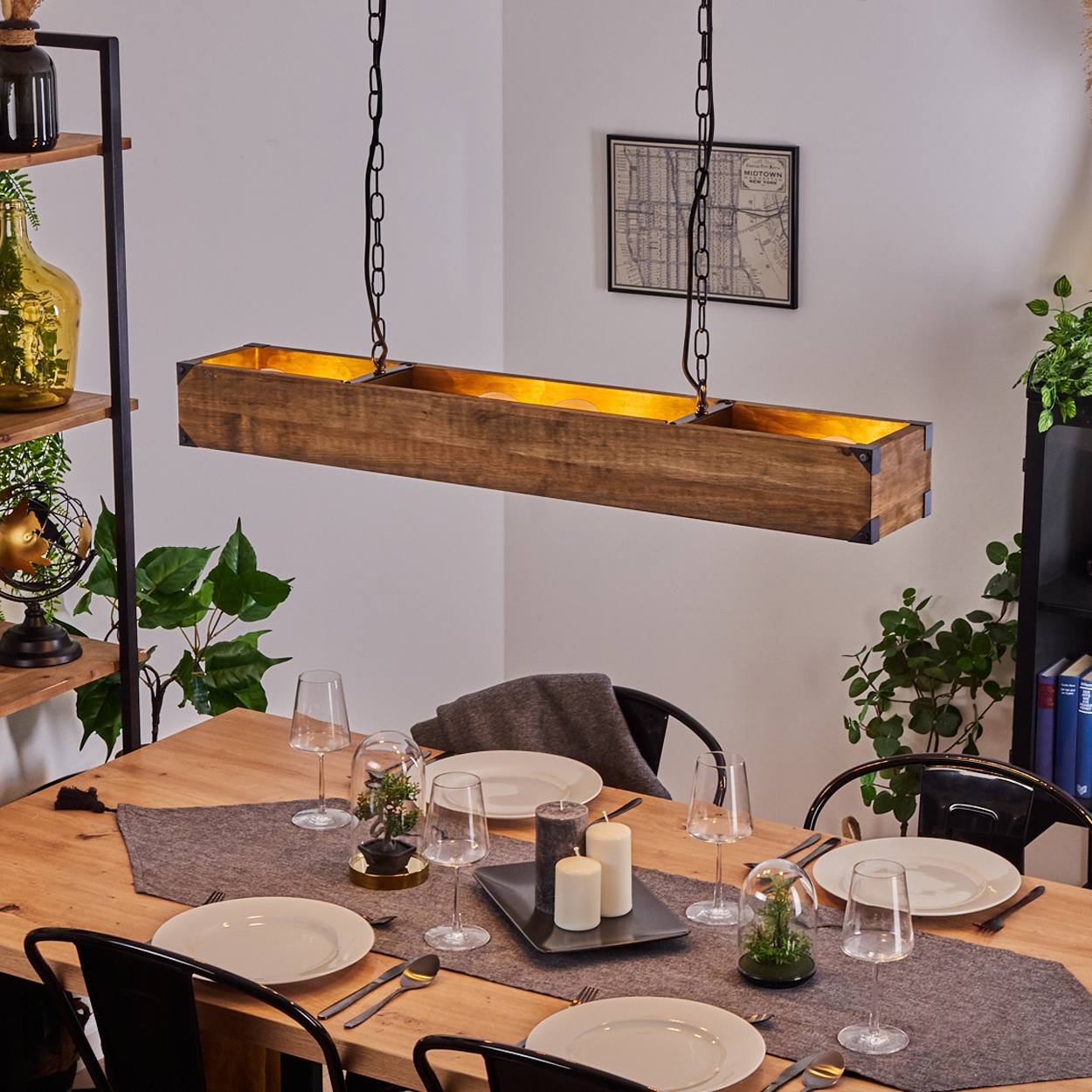 Ein gedeckter Esstisch. Eine rechteckige Holzkammer-Lampe hängt darüber. Teller, Gläser und Kerzen stehen auf dem Tisch. Regale mit Pflanzen an den Seiten.