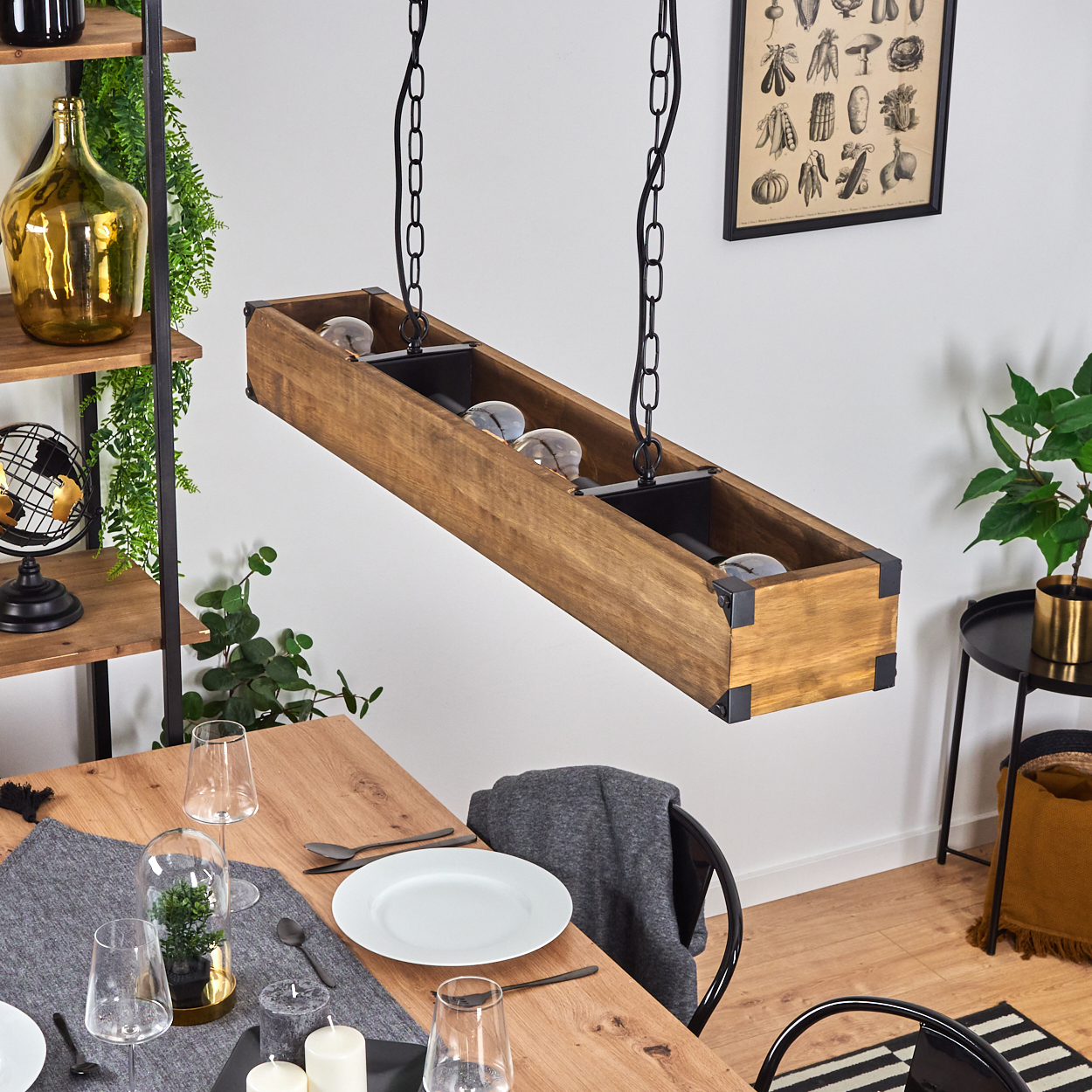 Hölzerne Leuchte mit Glühbirnen hängt über einem Tisch, der für das Abendessen gedeckt ist. Pflanzen und Dekorationsgegenstände sind im Hintergrund.