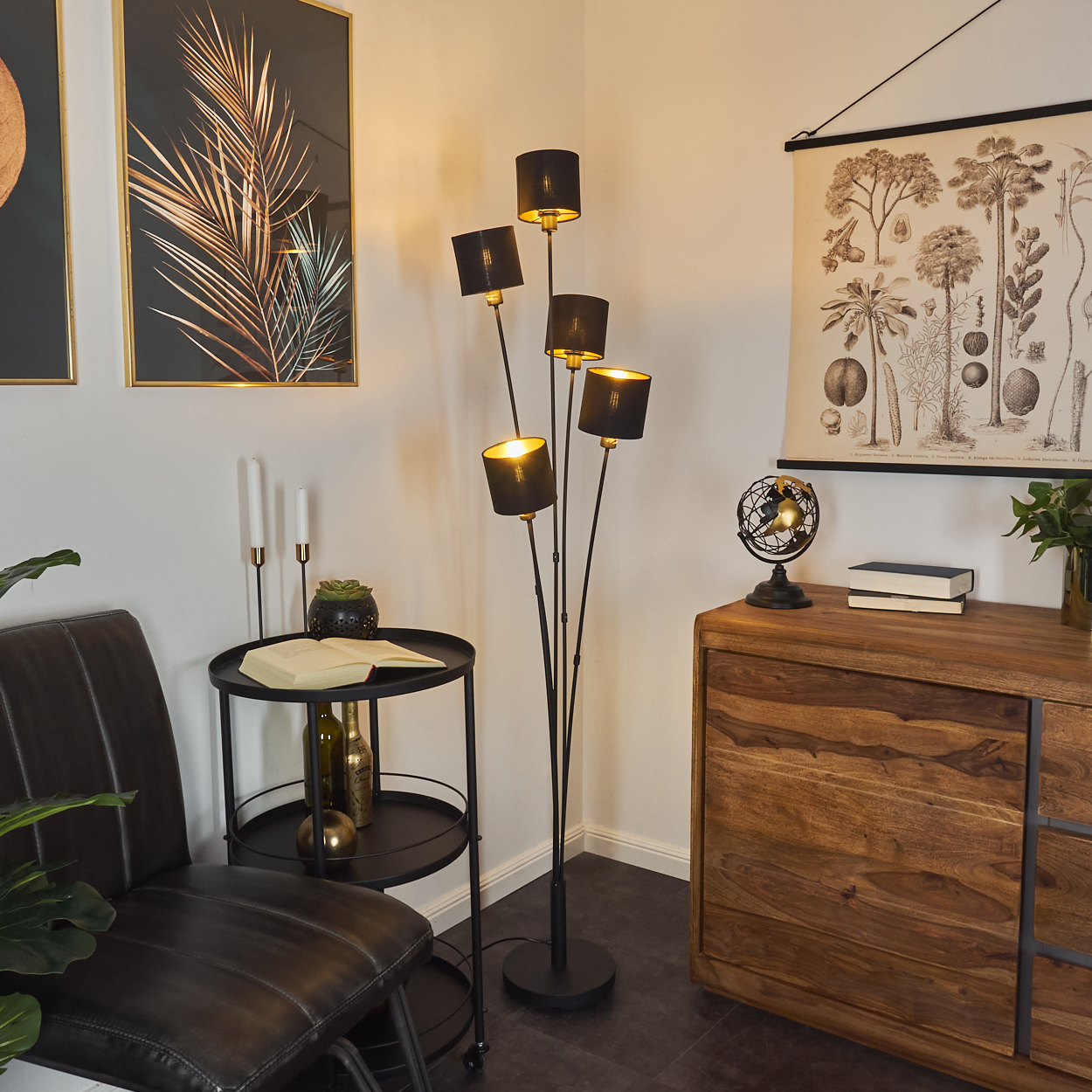 Ein Raum mit einer schwarzen Lampe, einem Holzschrank, einem schwarzen Beistelltisch mit Stuhl und botanischen Drucken an der Wand.