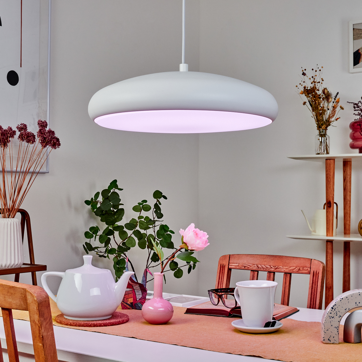 Ein Esstisch mit weißer Teekanne, Tasse und Blumenvase unter einer modernen Lampe. Rosa- und Brauntöne.
