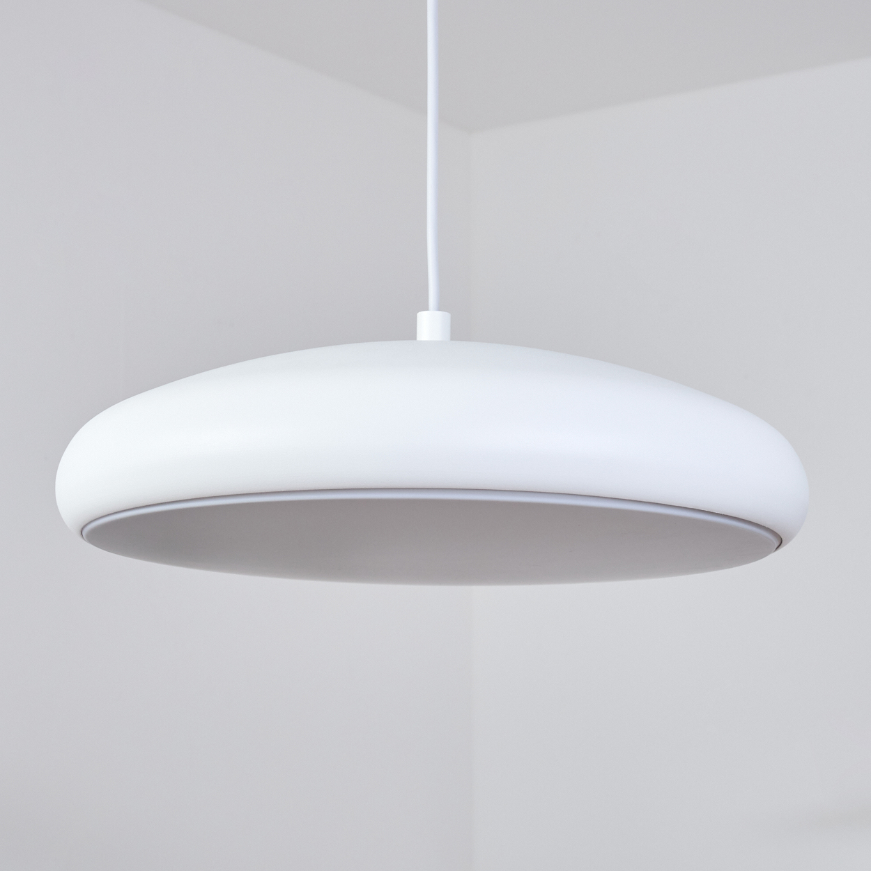 Eine weiße, moderne, scheibenförmige Deckenlampe hängt an einem weißen Kabel. Der Hintergrund ist eine weiße Zimmerecke.