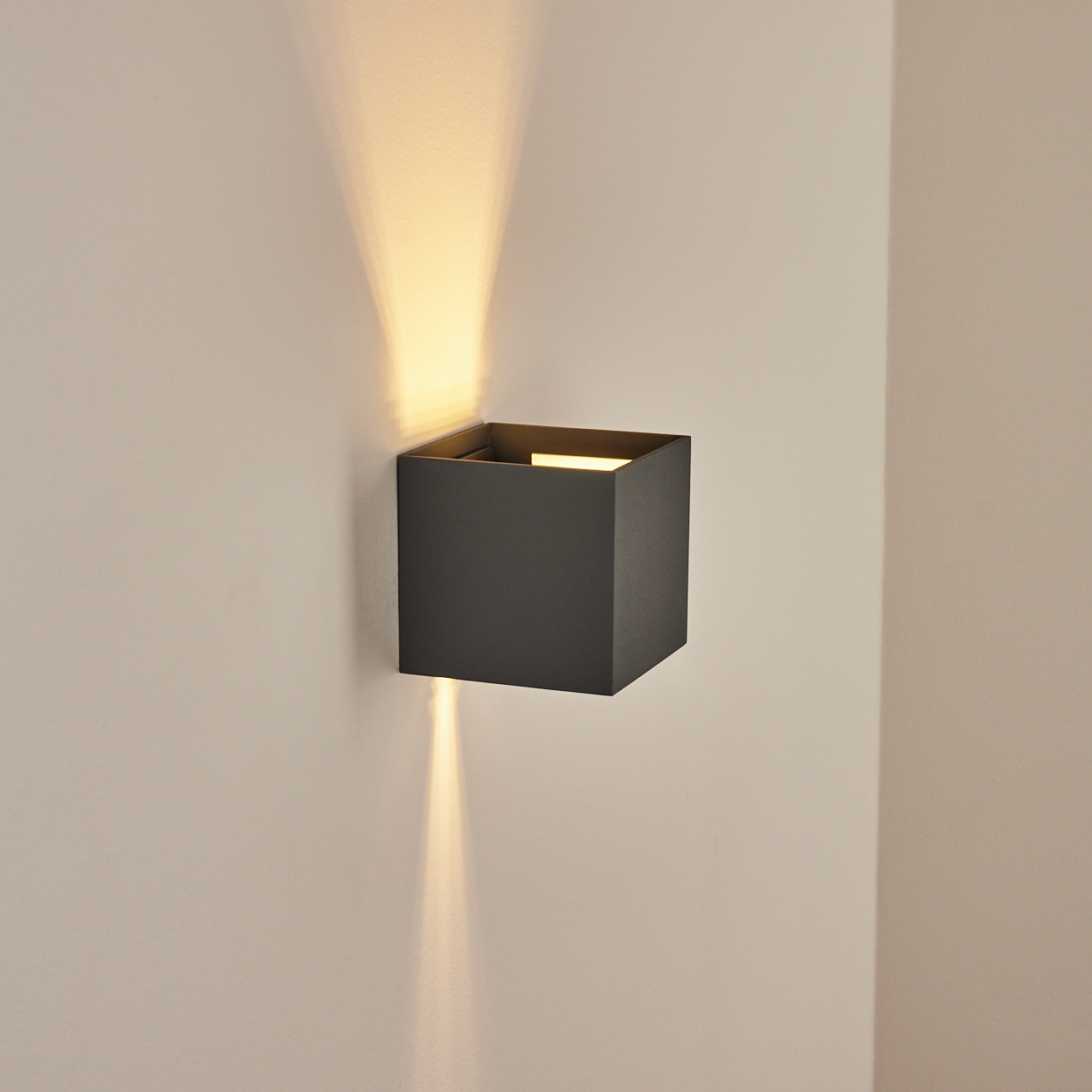 Eine schwarze, quadratische Wandleuchte strahlt Licht nach oben und unten aus und erzeugt ein Muster an einer beigen Wand.