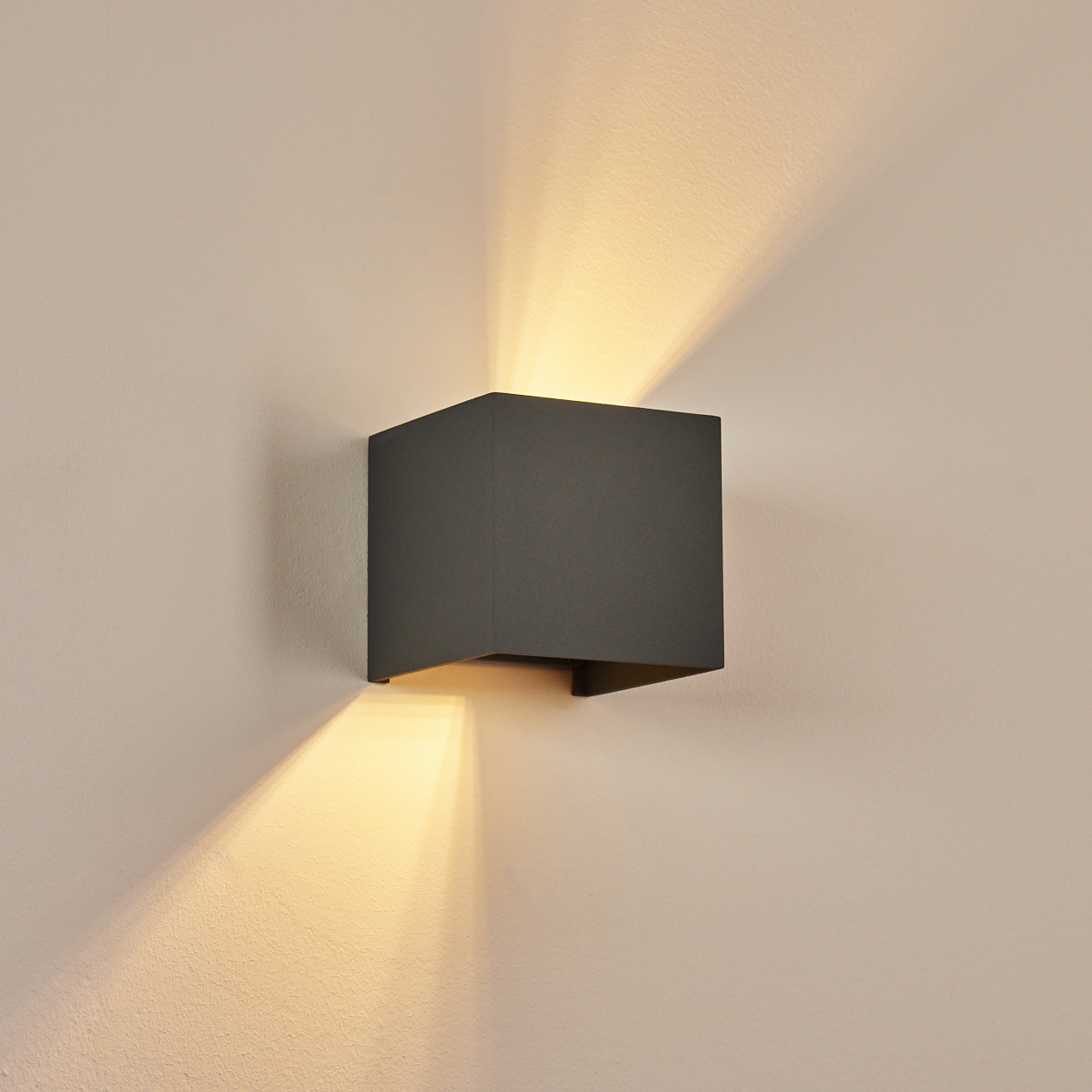 Eine dunkelgraue, würfelförmige Wandleuchte ist an einer beigefarbenen Wand montiert. Licht strahlt nach oben und unten. Die Wand hat eine strukturierte Oberfläche.