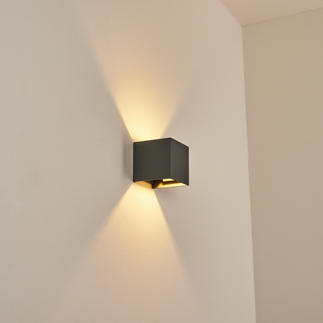 Eine quadratische, dunkelgraue Wandleuchte strahlt Licht nach oben und unten auf eine hellbeige Wand. Die Szene ist eine Ecke eines Raumes.