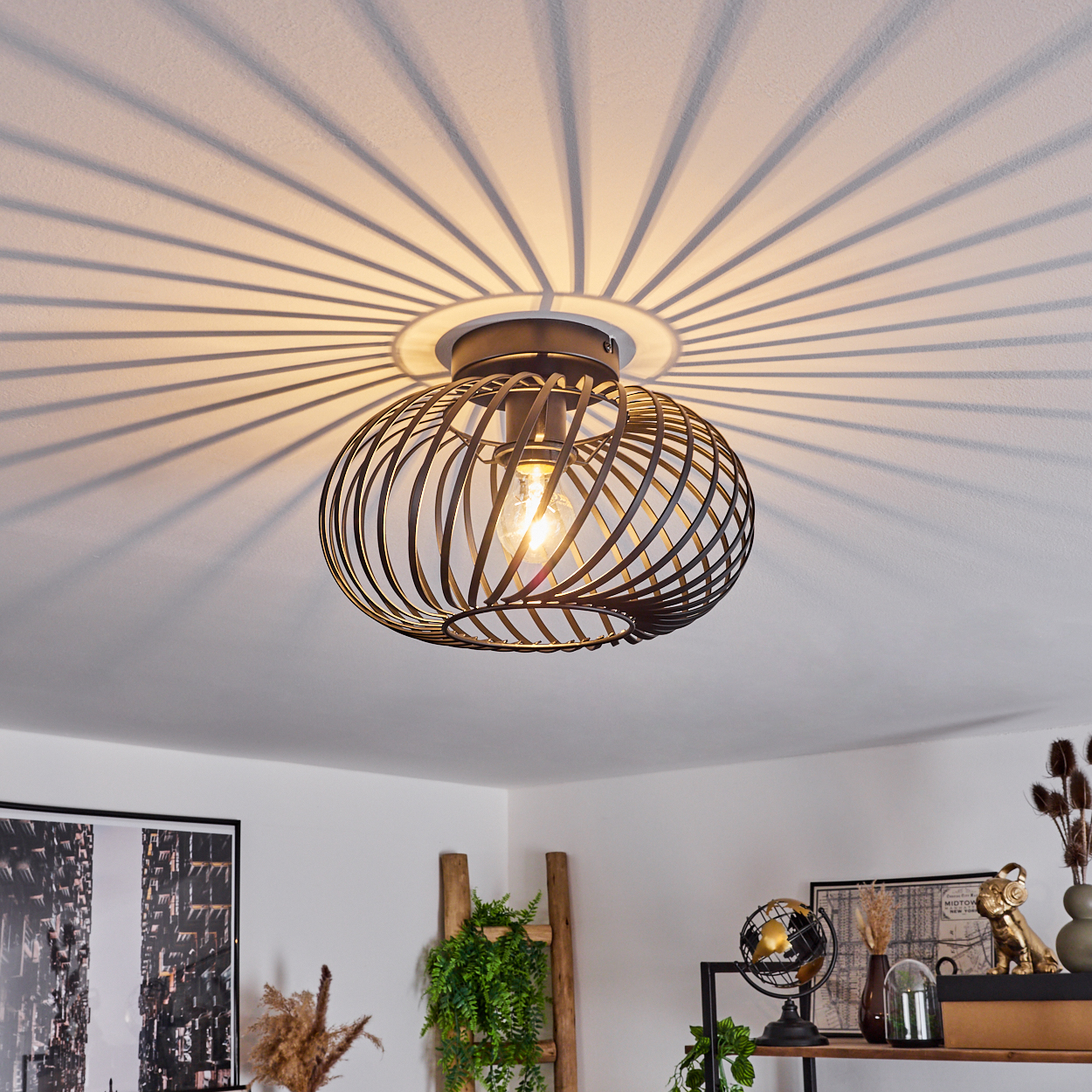 Eine Deckenlampe mit käfigartigem Design wirft radiale Schatten. Eine Glühbirne ist sichtbar. Deko-Artikel und eine Leiter im Hintergrund.