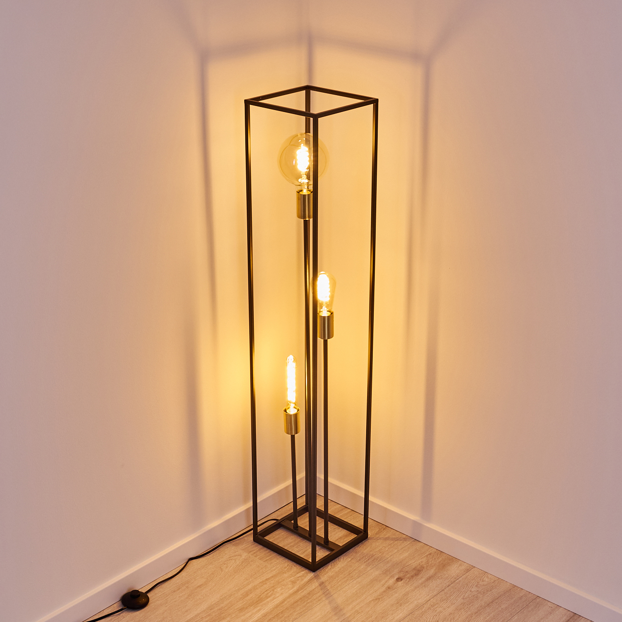 Eine schwarze Metall-Stehlampe mit drei leuchtenden Glühbirnen steht in einer Ecke. Die Wand und der Boden sind hell. Schatten.