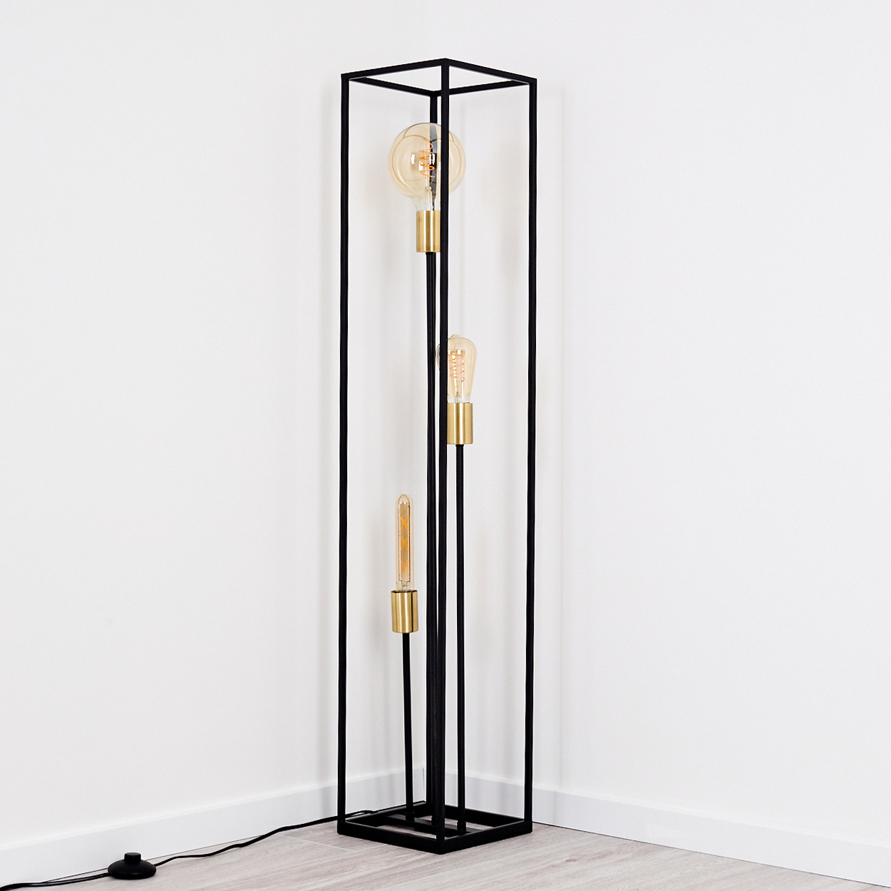 Eine schwarze Metall-Stehlampe mit drei Glühbirnen. Sie steht in einer Ecke eines Raumes mit weißen Wänden und Holzboden.