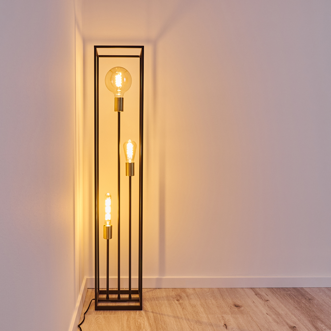 Eine schwarze, rechteckige Stehlampe mit drei leuchtenden Glühbirnen steht in einer Ecke. Holzboden, weiße Wände. Licht beleuchtet die Wand.