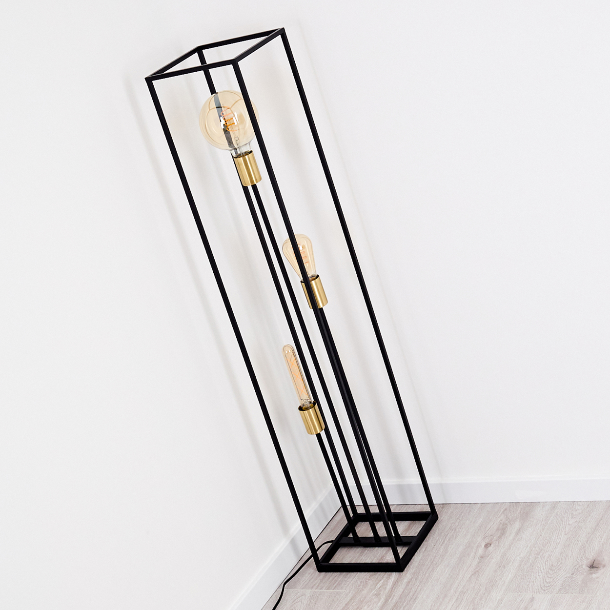 Eine schwarze Metall-Stehlampe mit drei freiliegenden Glühbirnen. Sie lehnt an einer weißen Wand. Der Boden ist aus Holz.