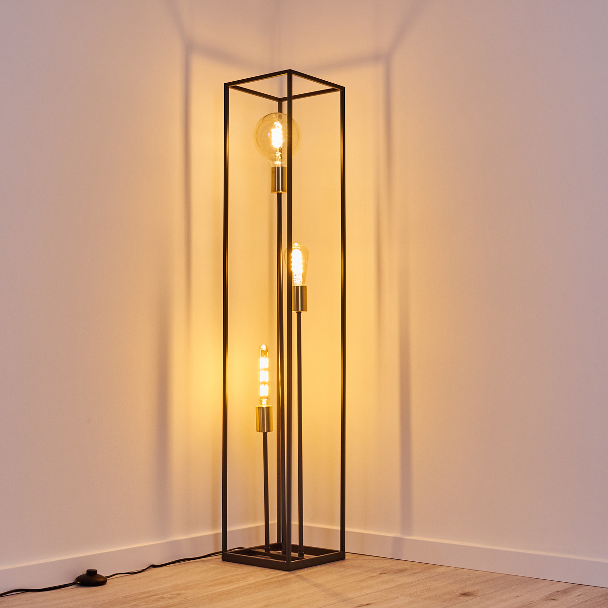 Eine schwarze Metall-Stehlampe mit drei brennenden Glühbirnen steht in einer Ecke. Die Wand ist weiß, der Boden helles Holz.