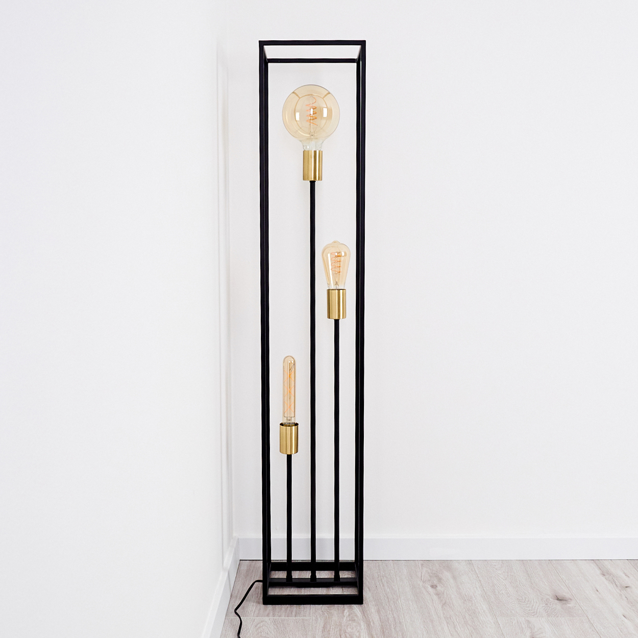 Eine moderne schwarze Stehlampe mit drei freiliegenden Glühbirnen. Die Glühbirnen haben unterschiedliche Formen und Größen, mit goldenem Sockel und Rahmen.